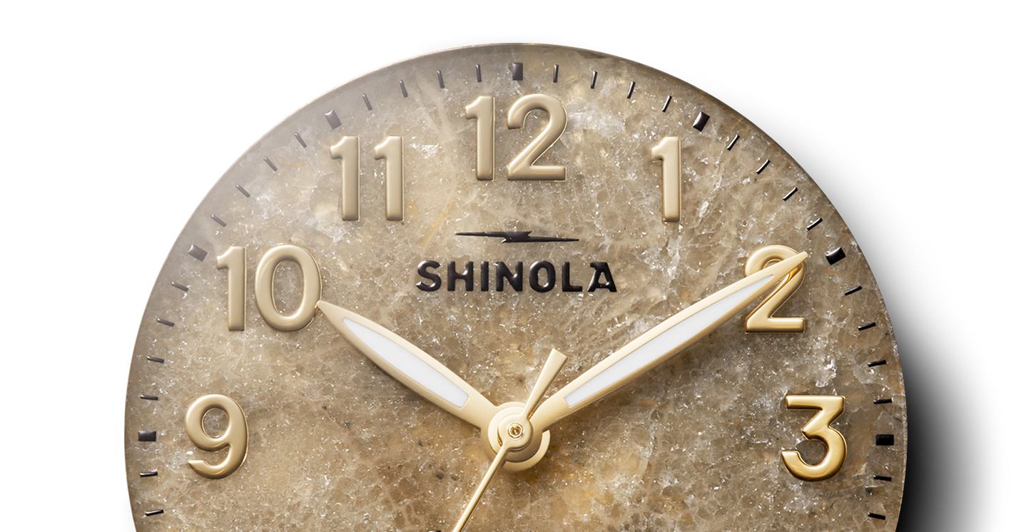 A close-up of the Shinola Birdy Watch - Petoskey, featuring a textured brownish face with gold numbers and white and gold hands, displays the time as 10:10:27.
