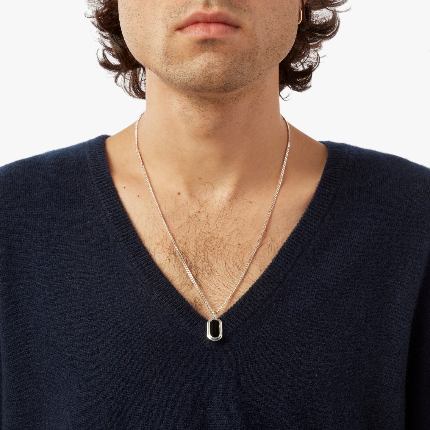A person, shown from shoulders to chin against a plain background, wears a dark blue V-neck sweater and the Shinola Onyx Tag Necklace – Sterling Silver, featuring a rectangular black onyx pendant.