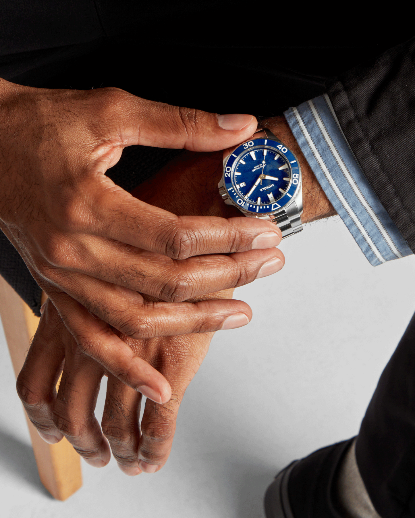 A close-up of a person’s wrist wearing the Shinola Lake Michigan Monster Automatic Dive Watch, featuring a stainless steel case and blue dial. Dressed in a dark suit and blue striped shirt, their hands rest over their lap.