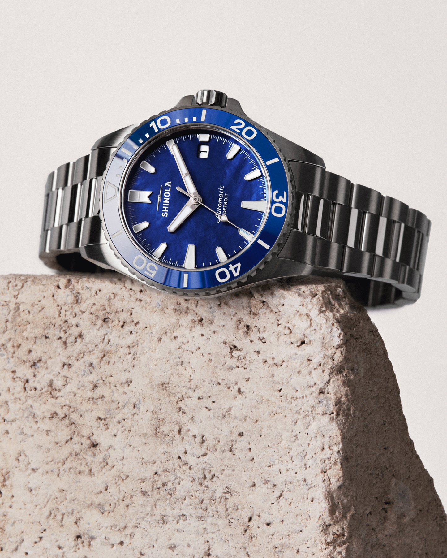 A Shinola Lake Michigan Monster Automatic Dive Watch with a blue face and bezel, silver stainless steel case, white hour markers, and an elegant bracelet rests on a rough, light-colored stone.