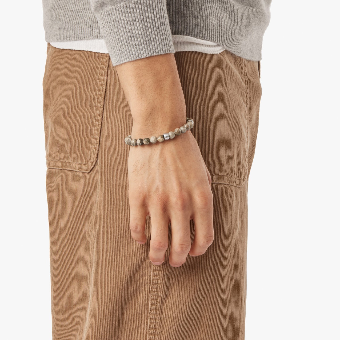 A person in light brown corduroy pants and a grey sweater wears the Shinola 8mm Beaded Petoskey Bracelet, shown on their wrist against a plain background, highlighting this unique Michigan jewelry accessory.
