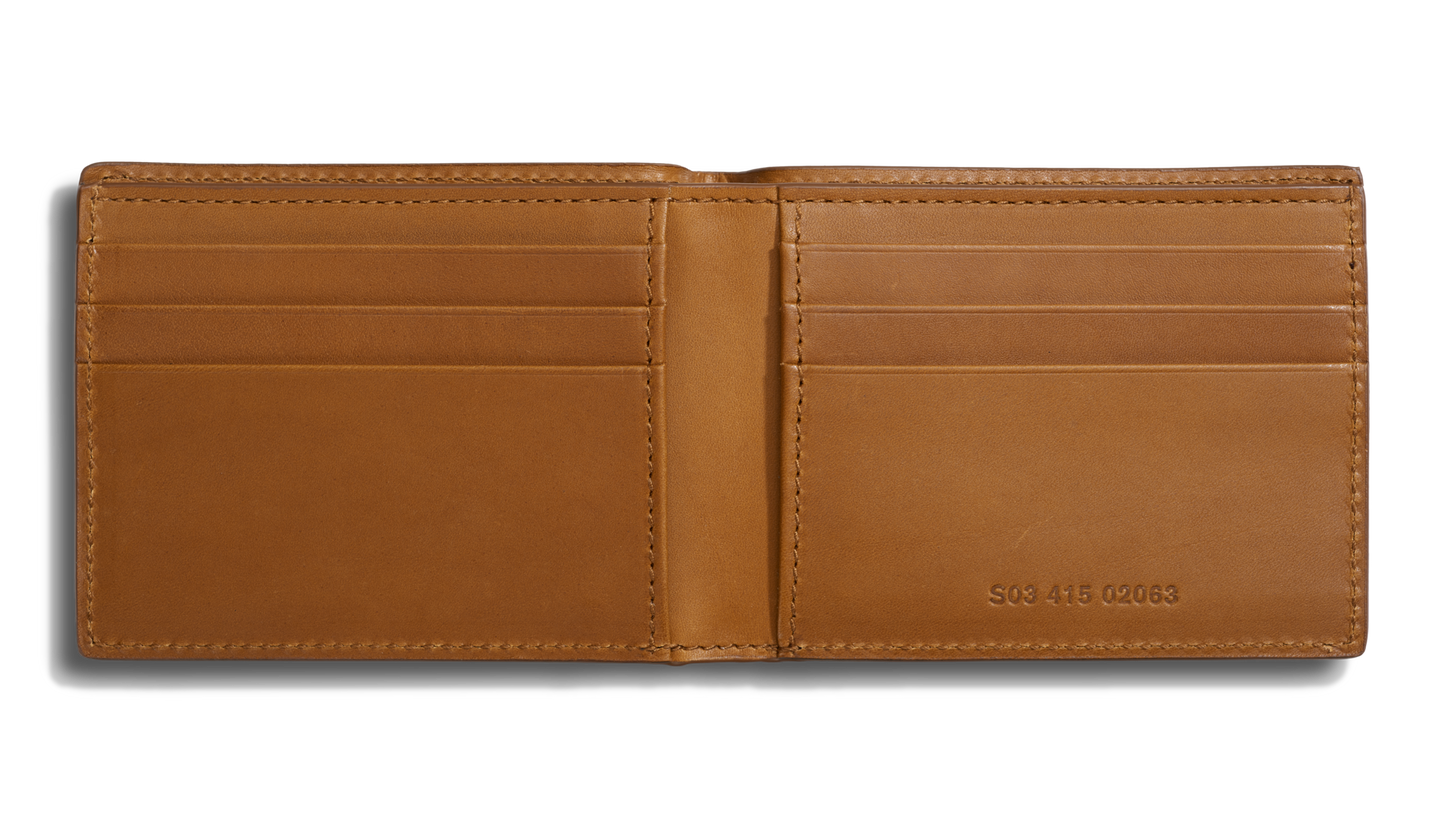 A tan Shinola Slim Bifold Wallet is open, revealing six card slots and stitched edges. S94 415 0063 is embossed on the lower right, adding detail to this stylish leather bifold wallet.