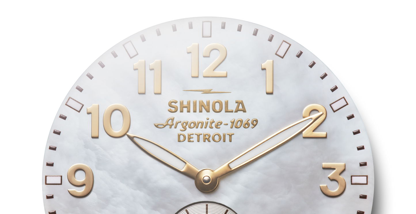 Close-up of a Shinola Runwell Watch - Mother Of Pearl showing 10:10, with gold-tone numbers and hands. The white marble-textured background complements its two-tone stainless steel bracelet.