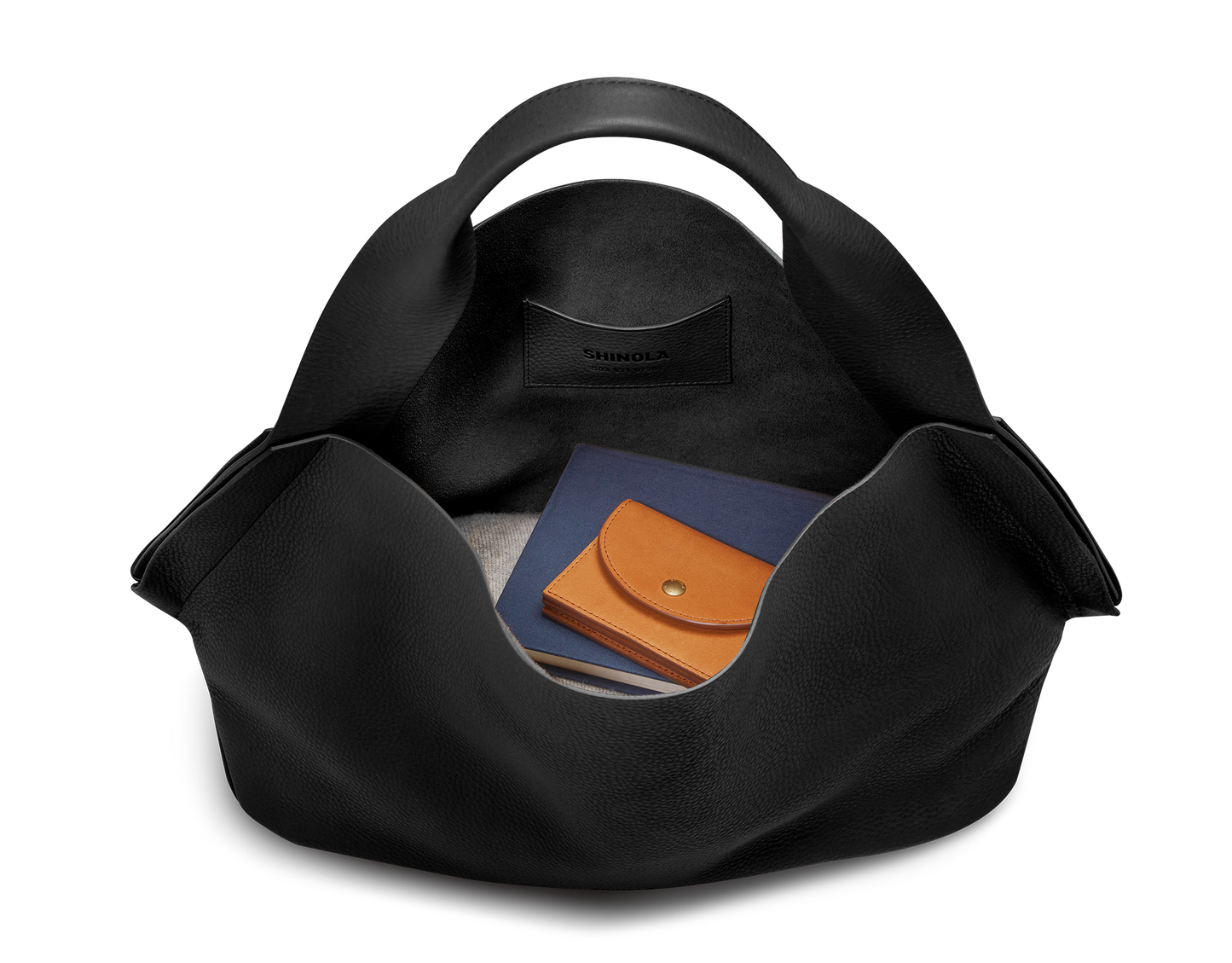 The Shinola Derby Shoulder Bag - Black has wide handles, a spacious open compartment with light fabric lining, and an interior leather card pocket. Inside are a blue notebook and tan leather wallet. The Shinola brand name appears on the card pocket.