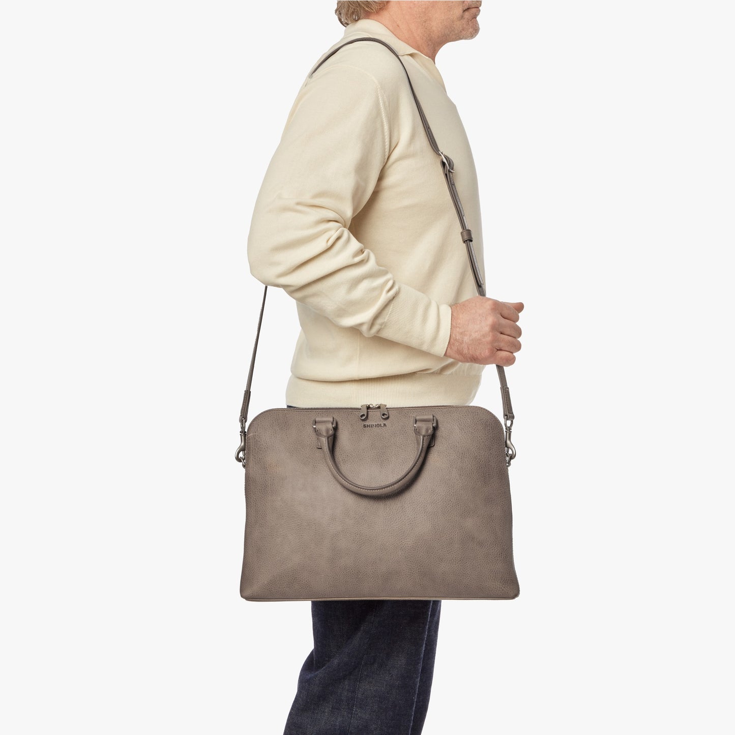 A person in a cream sweater and dark pants carries the Shinola Portfolio Briefcase - Stone, featuring handles and a shoulder strap, set against a plain white background.