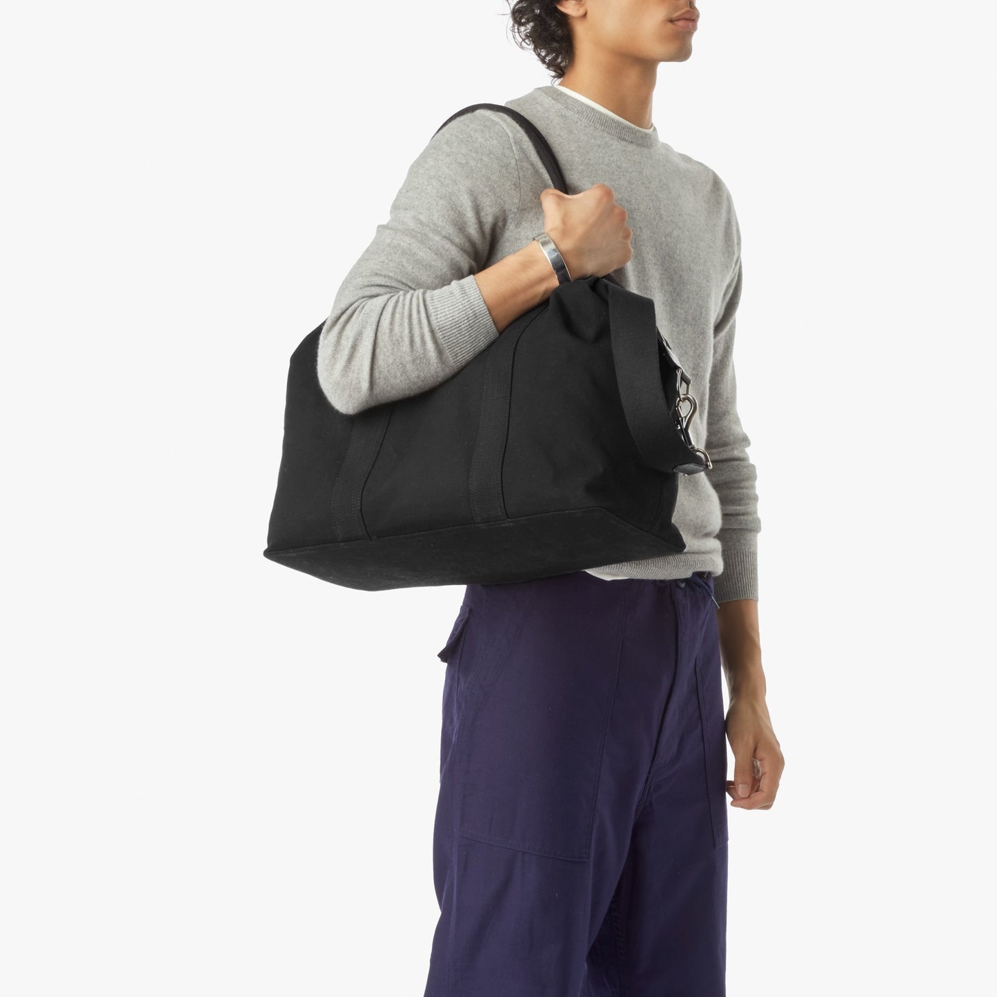 A person in a light grey sweater and blue pants holds the Shinola Runabout Holdall Canvas Bag in black over their shoulder, set against a plain white background. Their face is partially obscured.