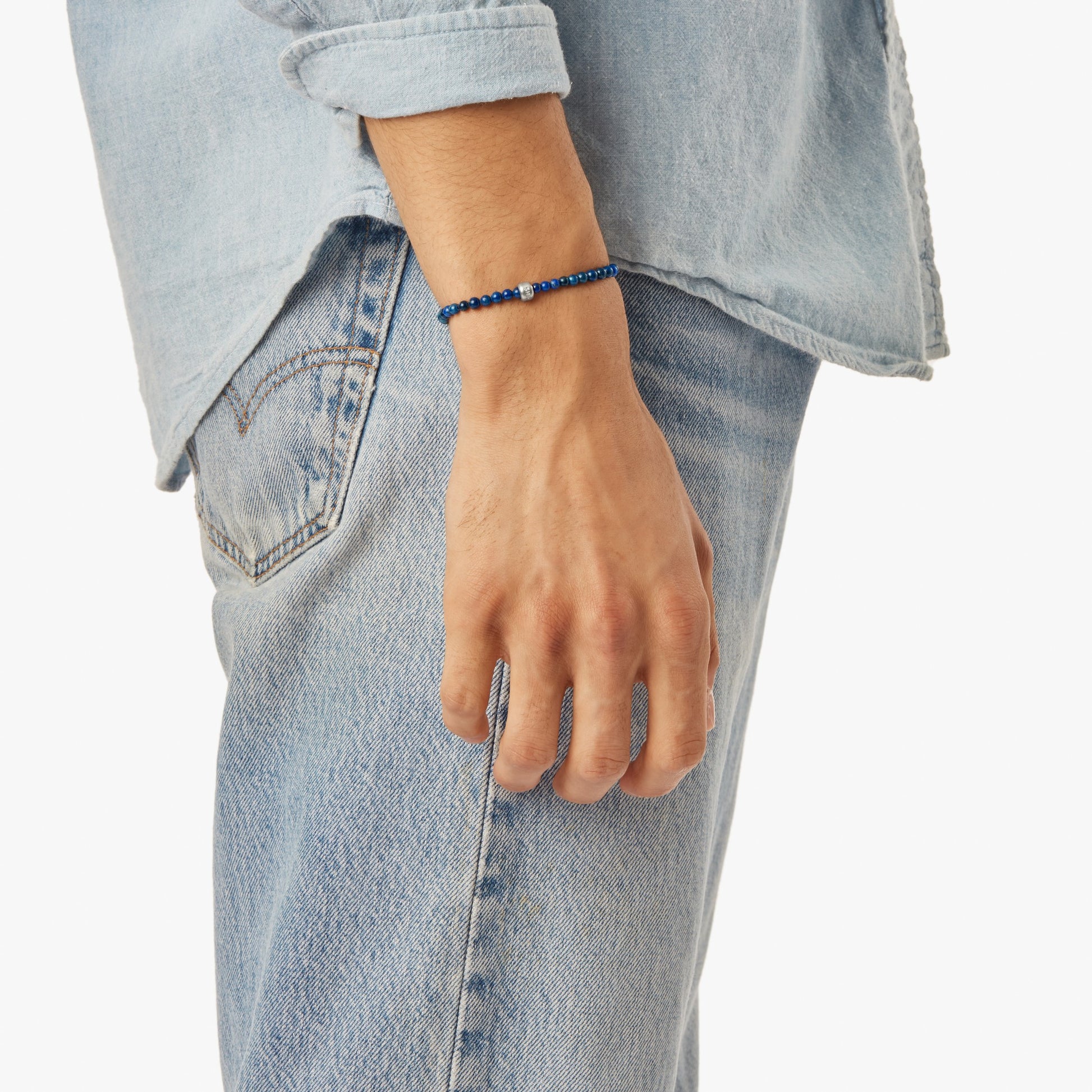 A person in a light blue denim shirt and jeans wears the Shinola 4mm Beaded Lapis Bracelet – Lapis, featuring lapis lazuli beads and a sterling silver accent, on their wrist with their arm relaxed by their side.