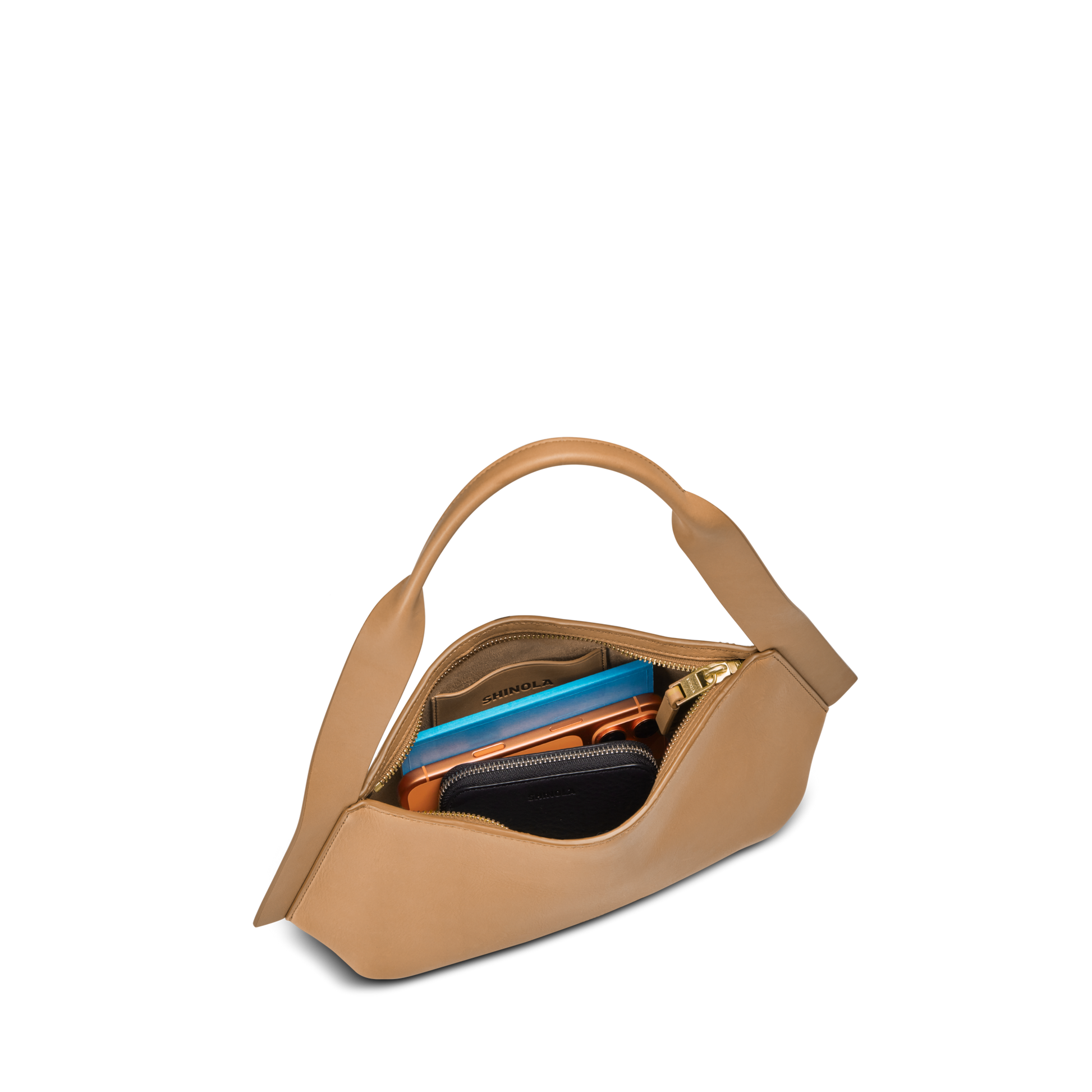 The Shinola Small Derby Shoulder Bag - Honey, a uniquely-shaped tan day-to-night bag with a top handle, sits open to reveal a blue notebook, zippered pouch, black wallet, and several small accessories neatly organized inside.