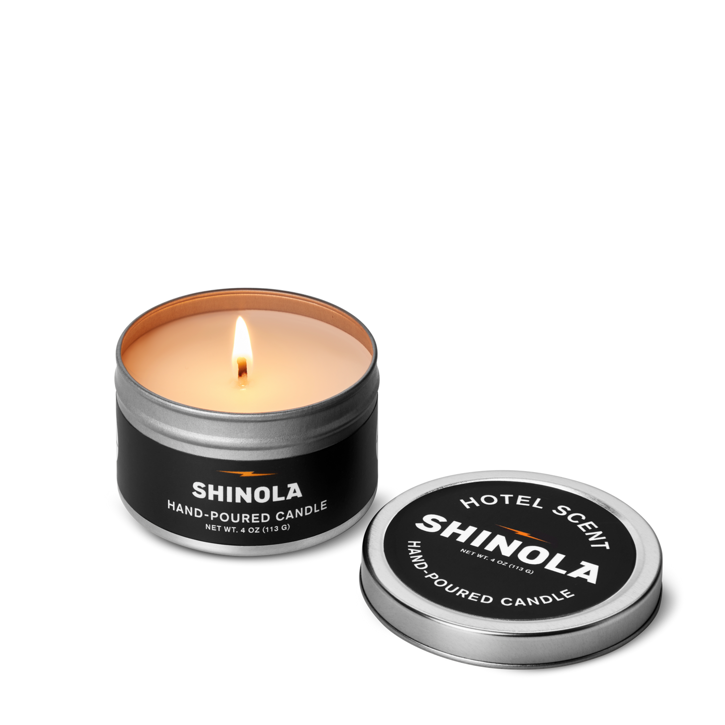 A lit Shinola Hand Poured 4oz Travel Candle - Hotel, made with coconut soy wax, glows in a sleek black tin with a silver rim and white text on both the tin and lid, making it a stylish portable scented candle.