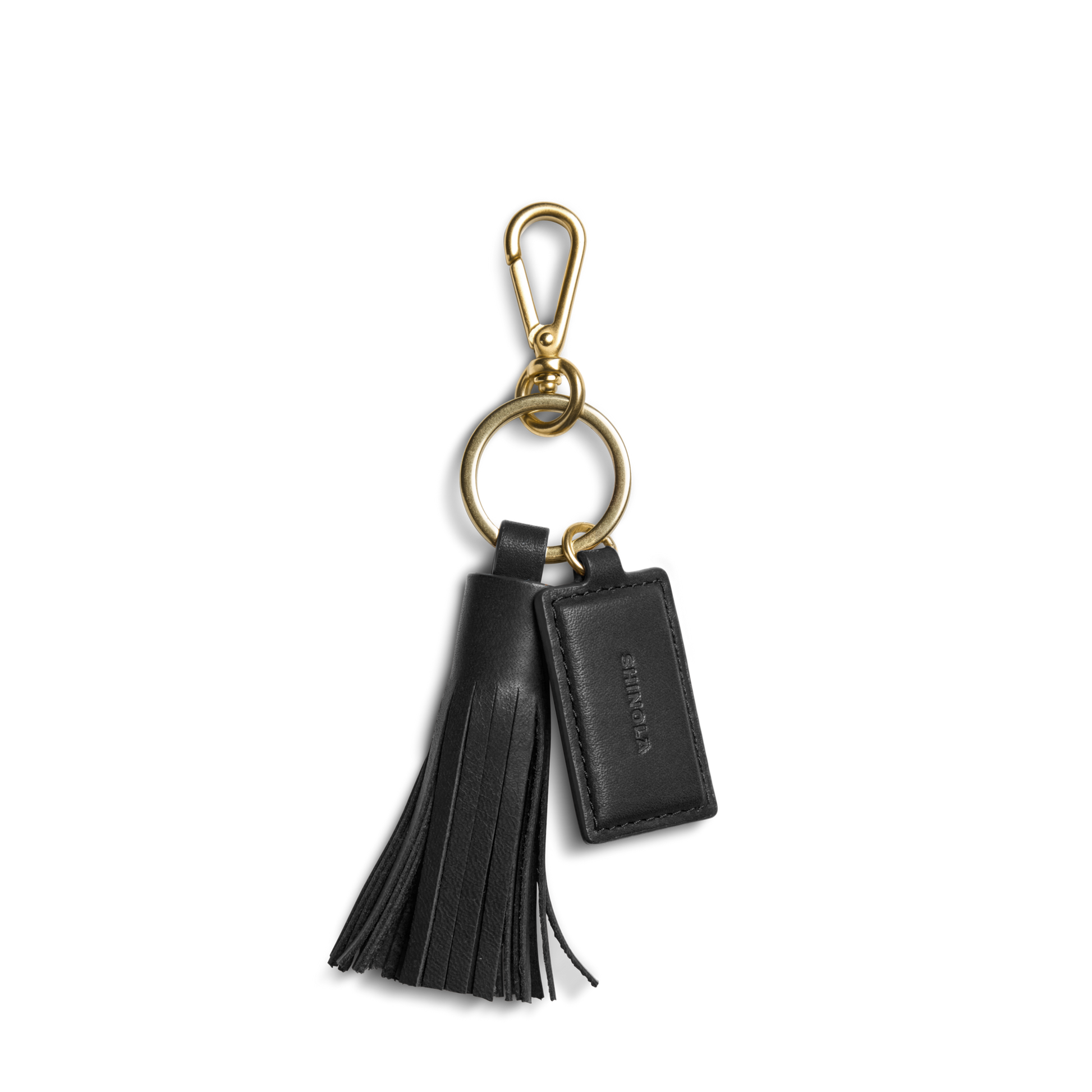The Shinola Leather Tassel Keychain - Black features a gold clasp and ring, a black leather tassel, and a rectangular embossed tag—an elegant monogram keychain or stylish key fob.