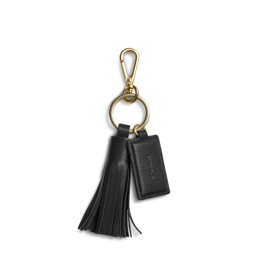 The Shinola Leather Tassel Keychain - Black features a gold clasp and ring, a black leather tassel, and a rectangular embossed tag—an elegant monogram keychain or stylish key fob.