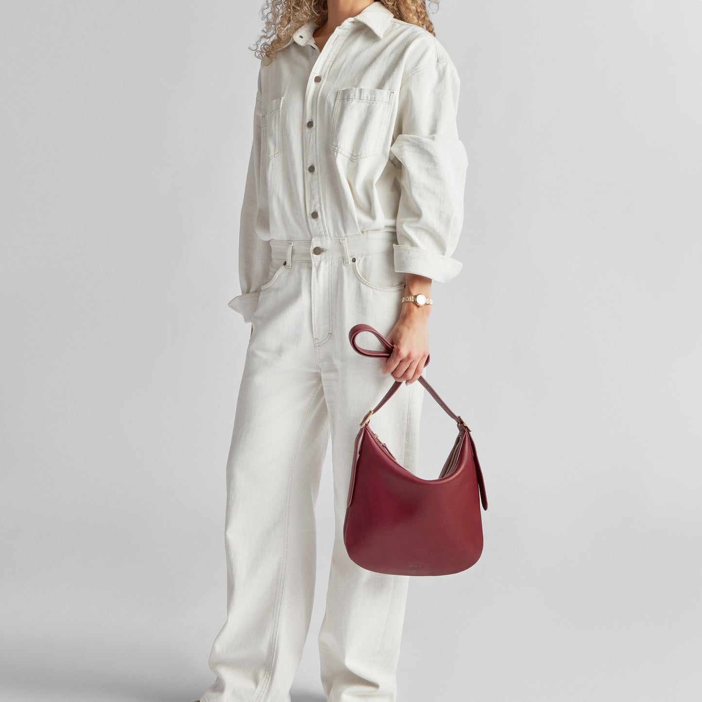A person in a white button-up jumpsuit stands against a plain background, holding the Shinola Medium Bixby Crossbody - Ruby in their left hand. Their face is not shown, and they have curly, light-colored hair.