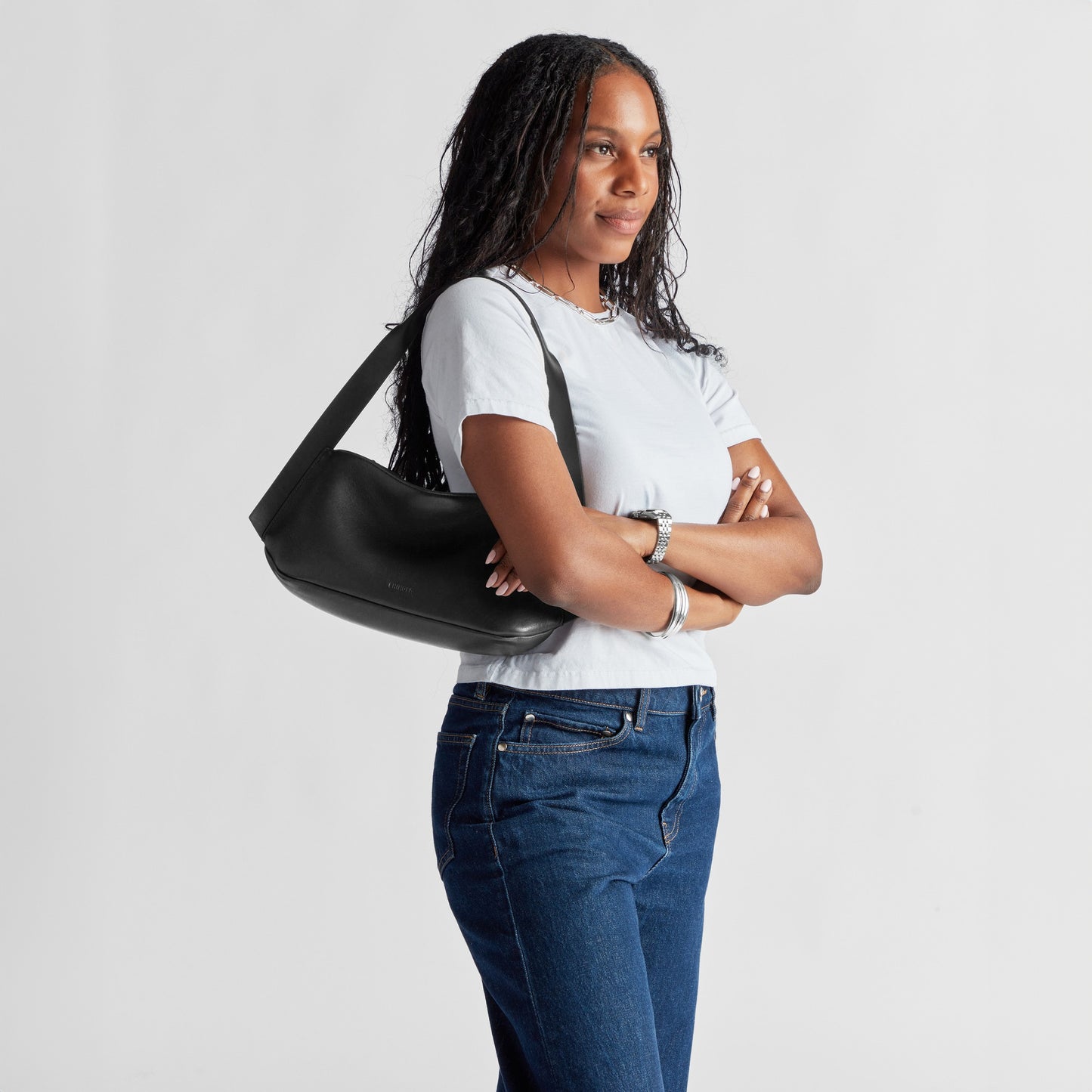 A Black woman with long, braided hair wears a white t-shirt and blue jeans, carrying the Shinola Small Derby Shoulder Bag in black natural leather across her body and accessorizing with silver jewelry on her neck and wrists.