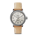 The Runwell Coin Edge Watch by Shinola features a beige leather strap, silver round case, white dial with silver Arabic numerals, three hands, and "Shinola" plus "Argonite-715 Detroit" printed above the center.
