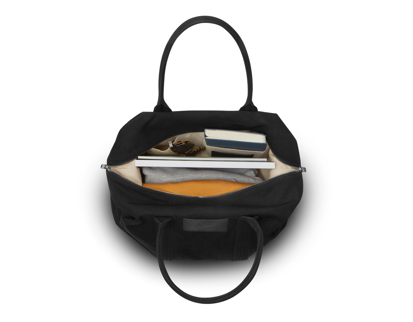 The Shinola Runabout Holdall Canvas Bag in black is pictured from above, revealing a beige interior neatly organizing a folded gray shirt, yellow item, glasses, two books, and a phone in separate compartments.