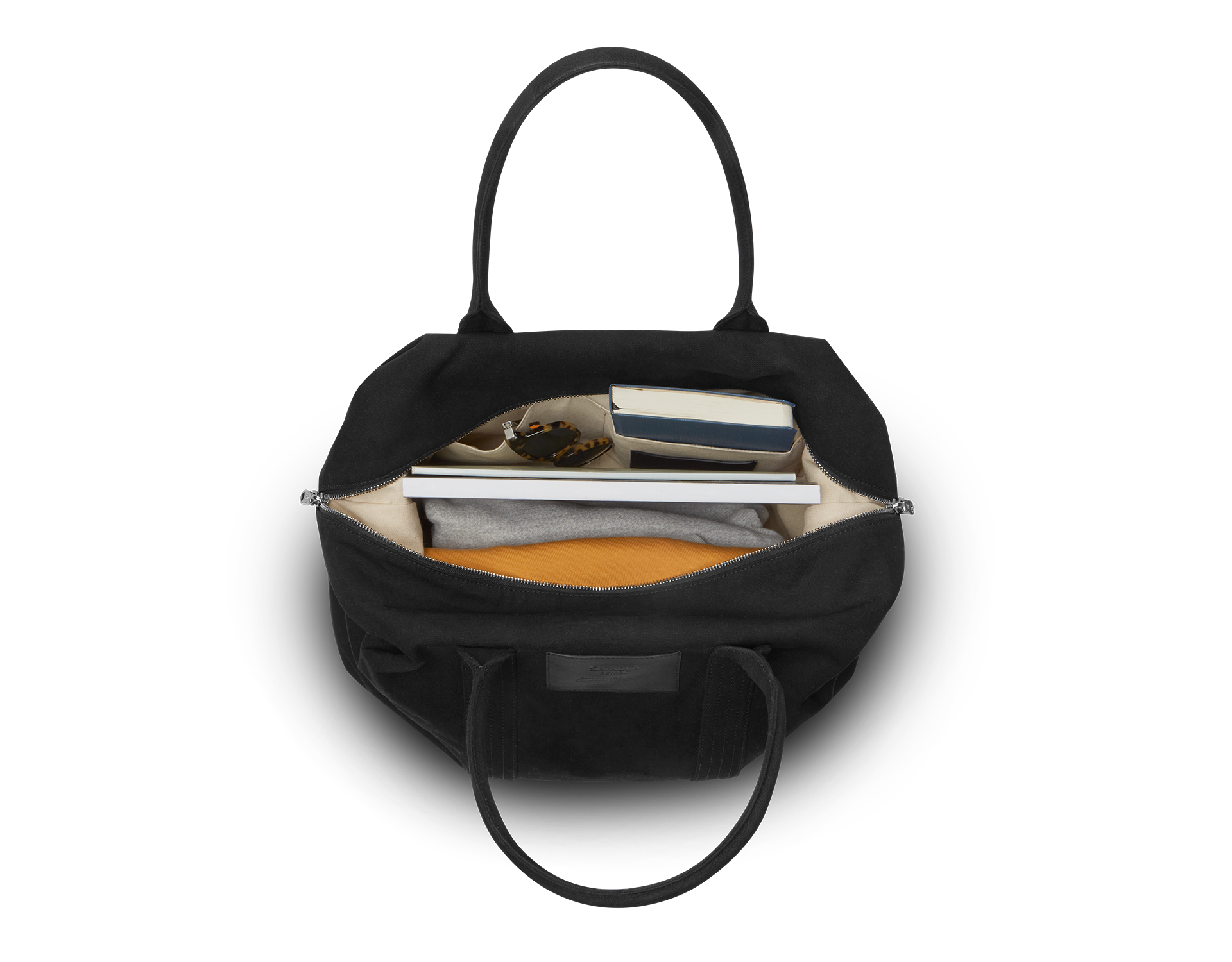 The Shinola Runabout Holdall Canvas Bag in black is pictured from above, revealing a beige interior neatly organizing a folded gray shirt, yellow item, glasses, two books, and a phone in separate compartments.