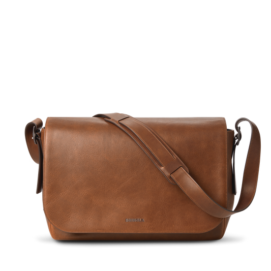 The Runwell Messenger Bag in medium brown by Shinola features a large flap closure, an adjustable crossbody strap, and the Shinola logo embossed near the bottom center. Displayed on a white background.