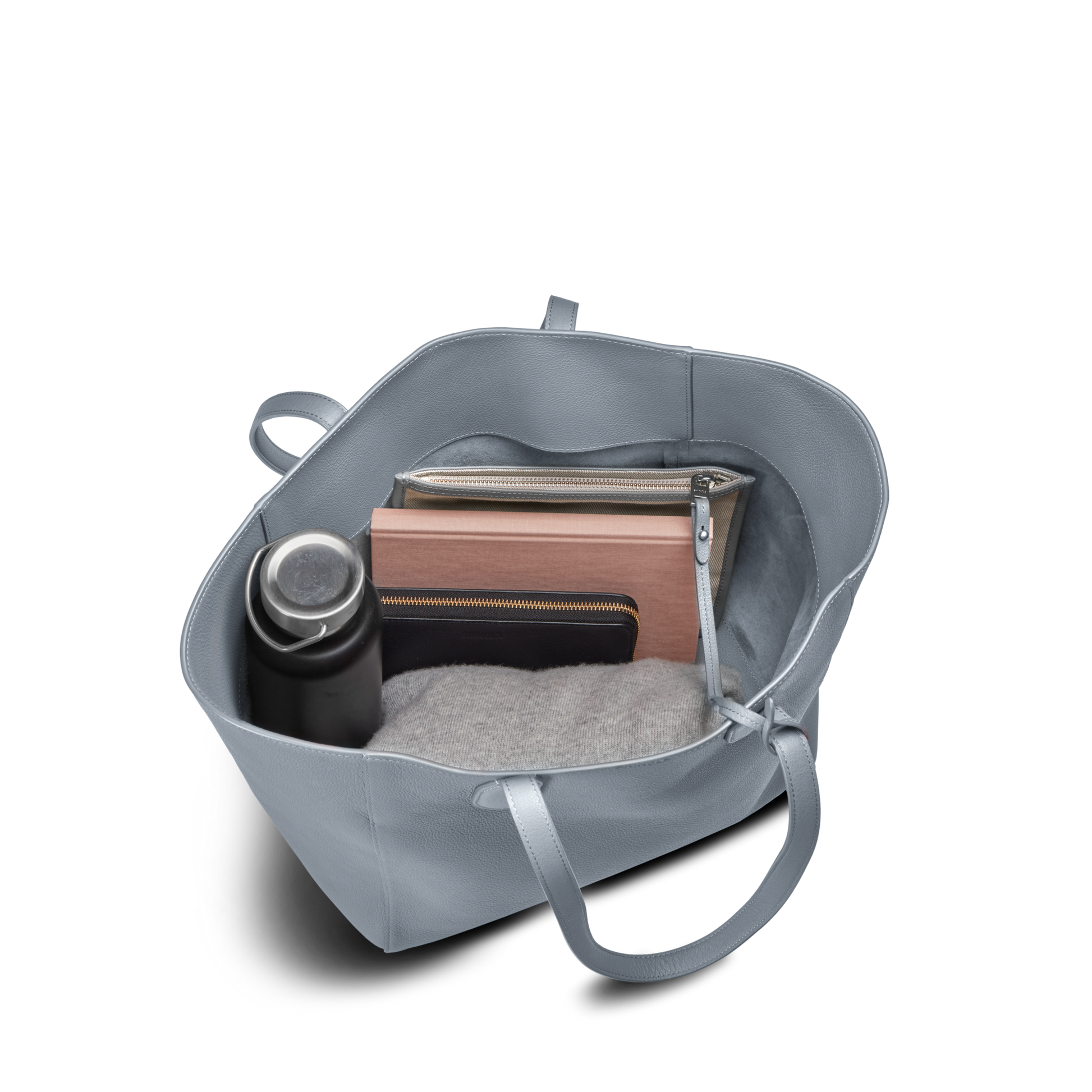 The Shinola Runabout Tote Bag - Mist is a light blue, lightweight tote shown from above, holding a water bottle, pebbled leather zipper pouch, closed laptop or tablet, notebook, and folded gray sweater.