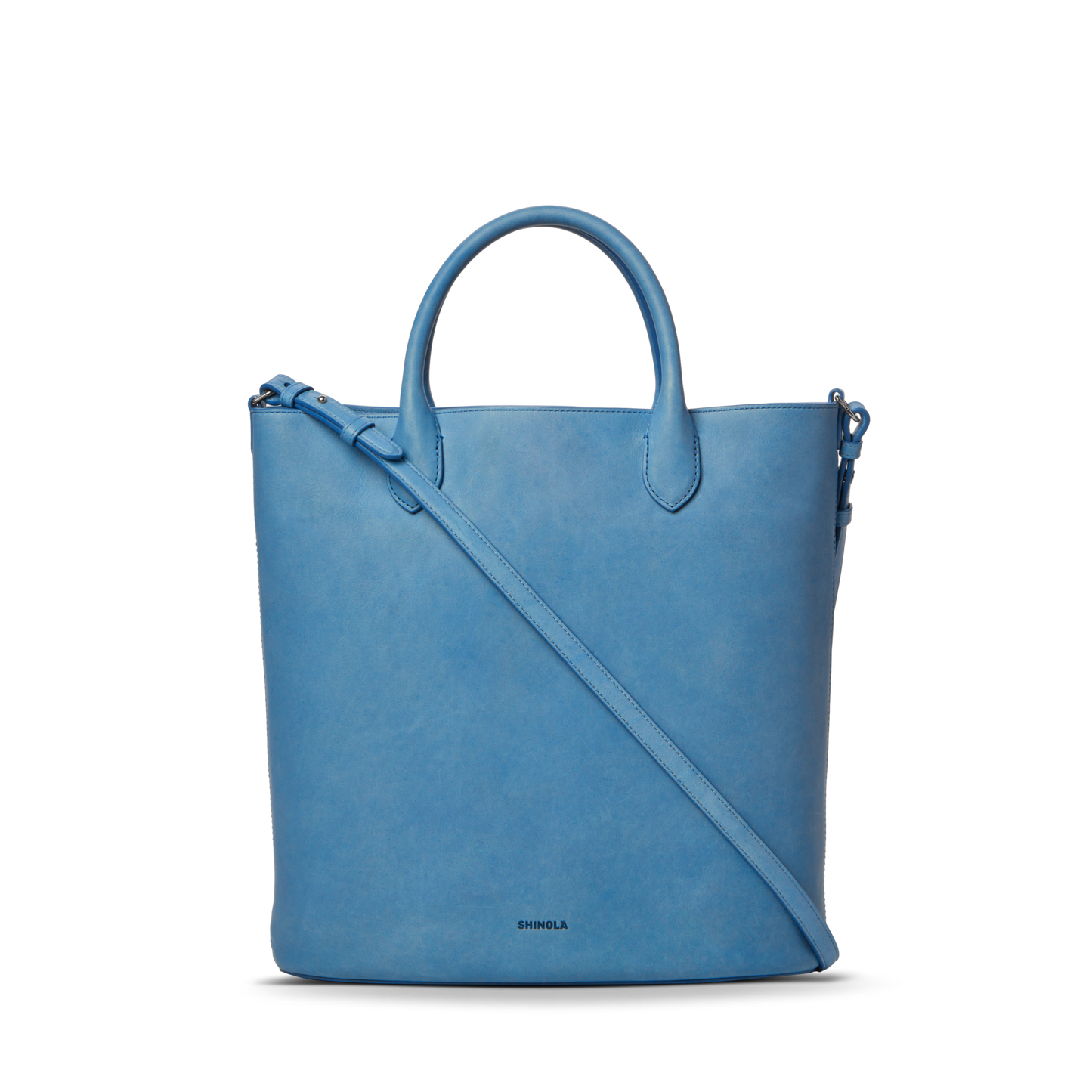 The Shinola 12 Hour Tote Bag - River is a light blue natural leather tote with two short handles, a long adjustable shoulder strap, minimal detailing, and the brand name printed near the bottom center.