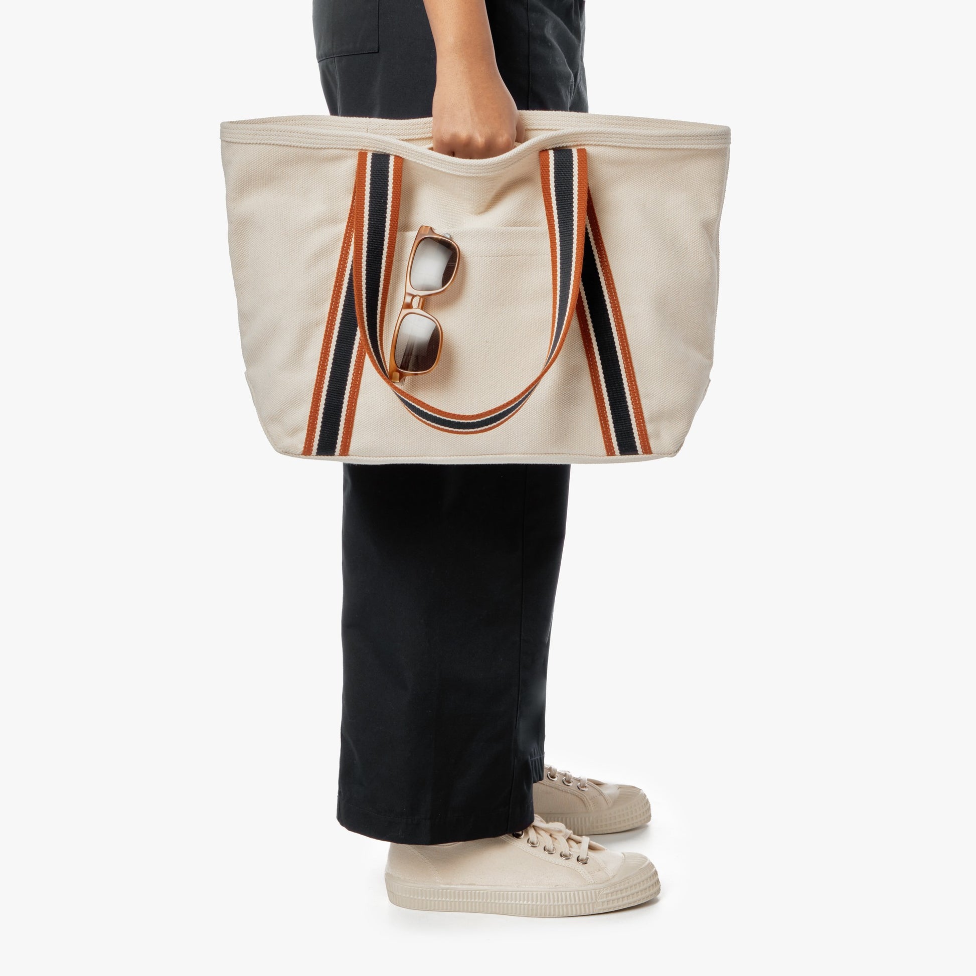 A person wearing black pants and beige sneakers holds the Shinola Small Easy Canvas Tote - Natural with striped handles and sunglasses on the strap—ideal for everyday essentials—while standing against a white background.