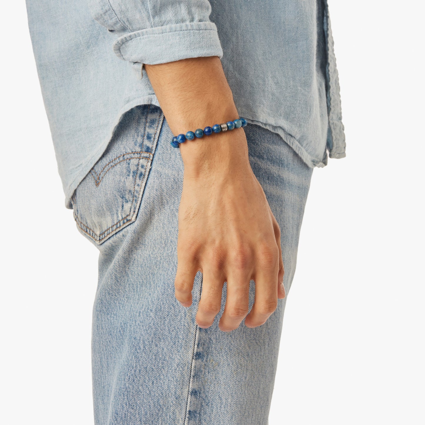 A person in a light denim shirt and jeans stands with their left arm relaxed, displaying the Shinola 8mm Beaded Lapis Bracelet - Lapis on their wrist against a white background.