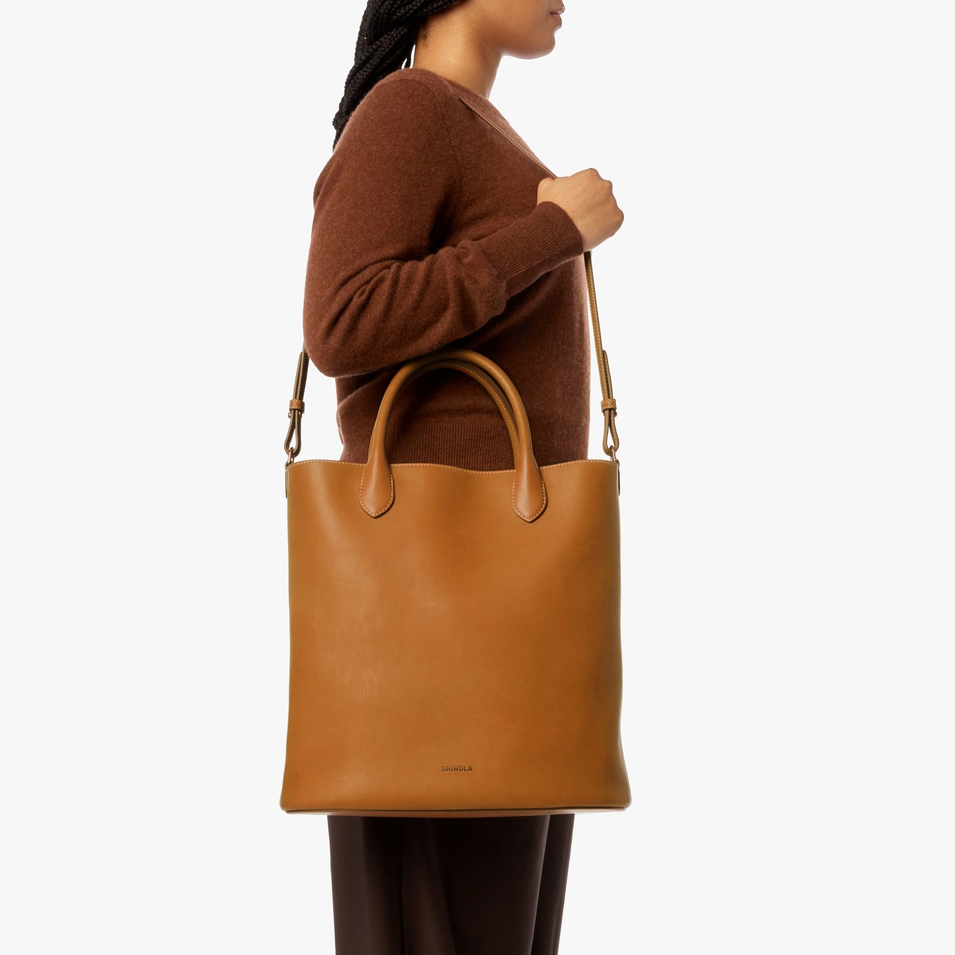 A person in a brown sweater and dark pants holds the Shinola 12 Hour Tote Bag - Tan, designed for daily essentials with both shoulder and hand straps, against a plain white background.