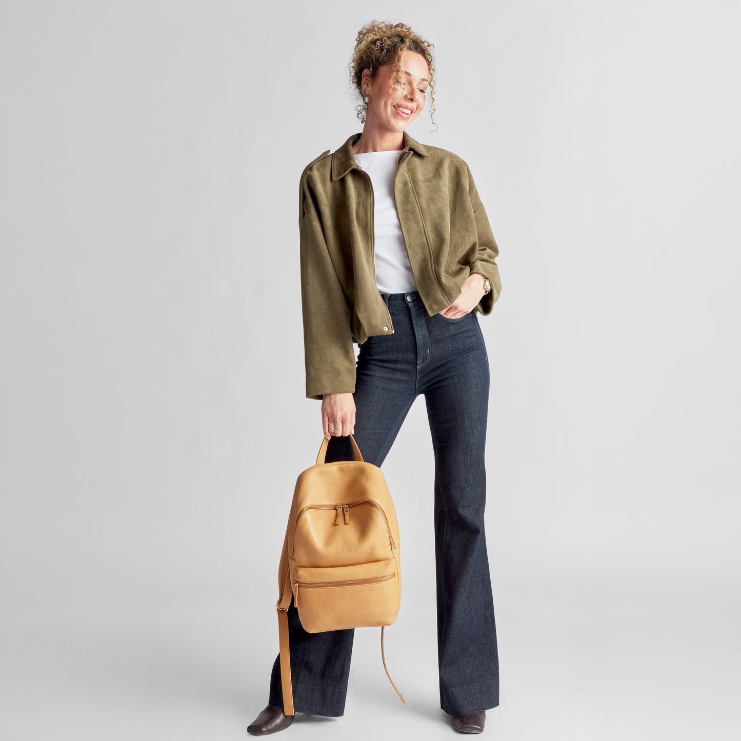 A woman smiles holding a Shinola Small Runwell Backpack in Honey, crafted from natural grain leather, with one hand. She wears a khaki jacket, white shirt, and dark flared jeans against a plain light gray background.