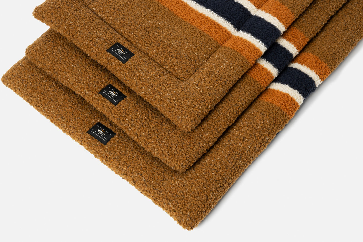 A stack of Shinola Crate Comfort Mat - Stripe Brown, cozy textured mats resembling shearling Berber dog beds with black labels and orange, white, and navy stripes near the edge, arranged neatly on a white background.
