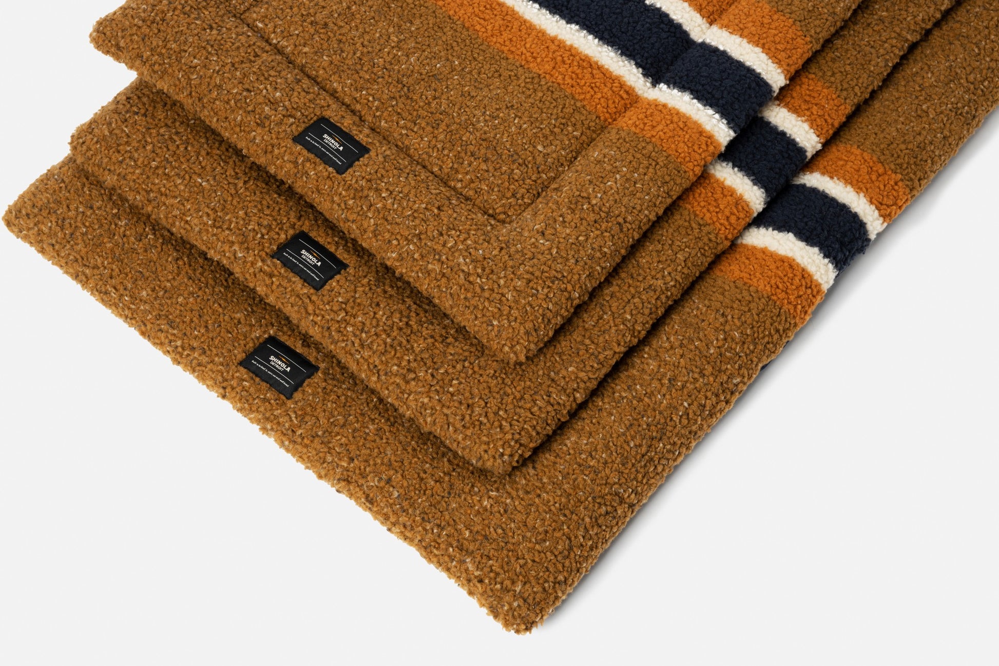 A stack of Shinola Crate Comfort Mat - Stripe Brown, cozy textured mats resembling shearling Berber dog beds with black labels and orange, white, and navy stripes near the edge, arranged neatly on a white background.