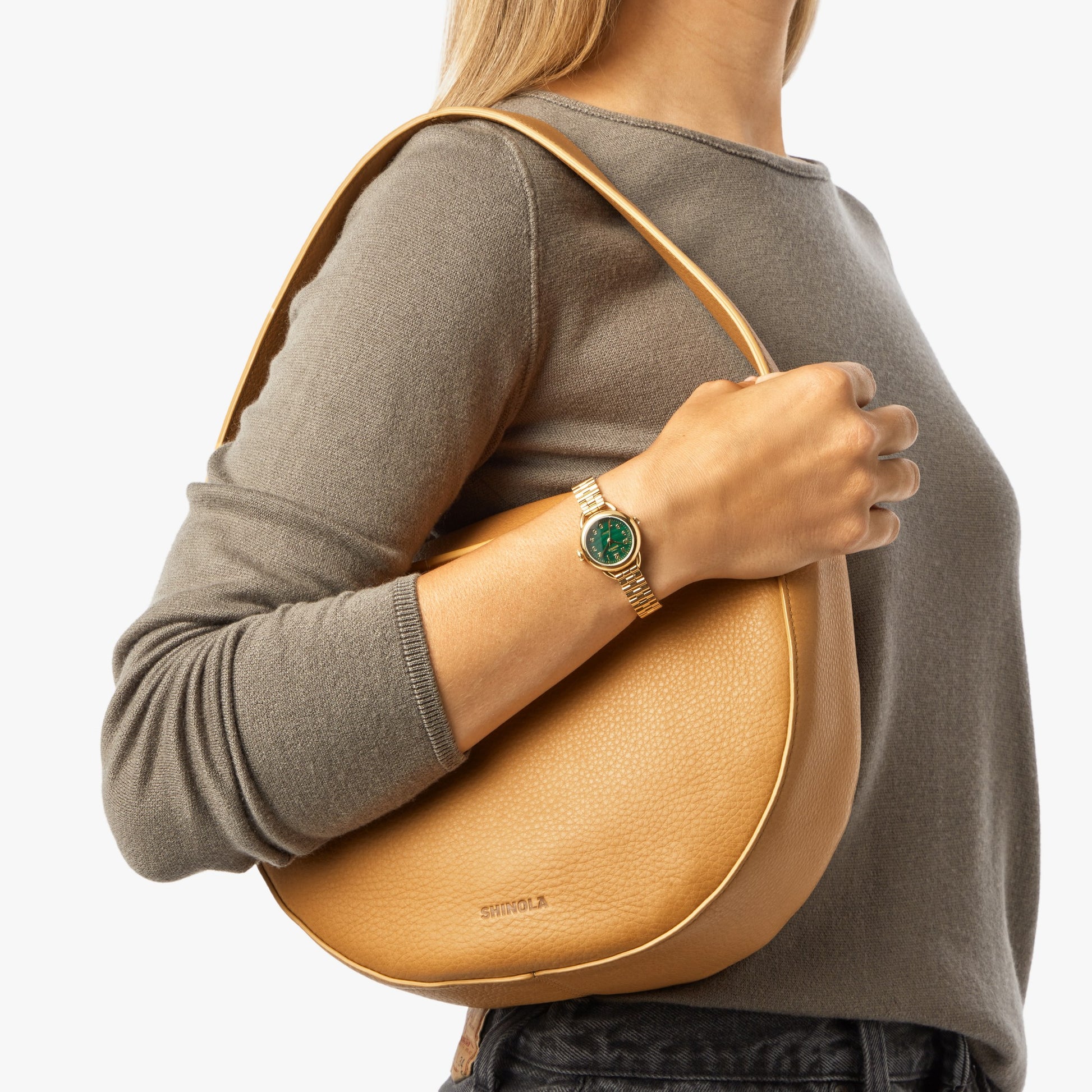 A person in a gray long-sleeve shirt holds the Shinola Small Shoulder Bag - Honey, a tan crescent-shaped leather bag, over their shoulder and displays a gold wristwatch with a green face. The background is white.