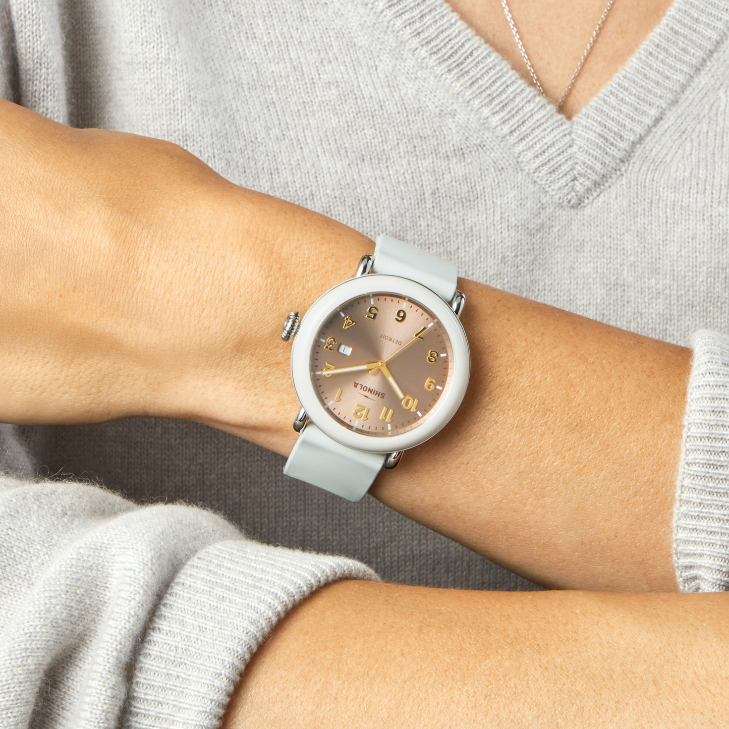 A person with crossed arms on their lap wears a light grey, long-sleeve sweater and displays the Shinola Detrola Watch - Nude, featuring a pale grey strap and rose gold face—a perfect showcase of classic American design.