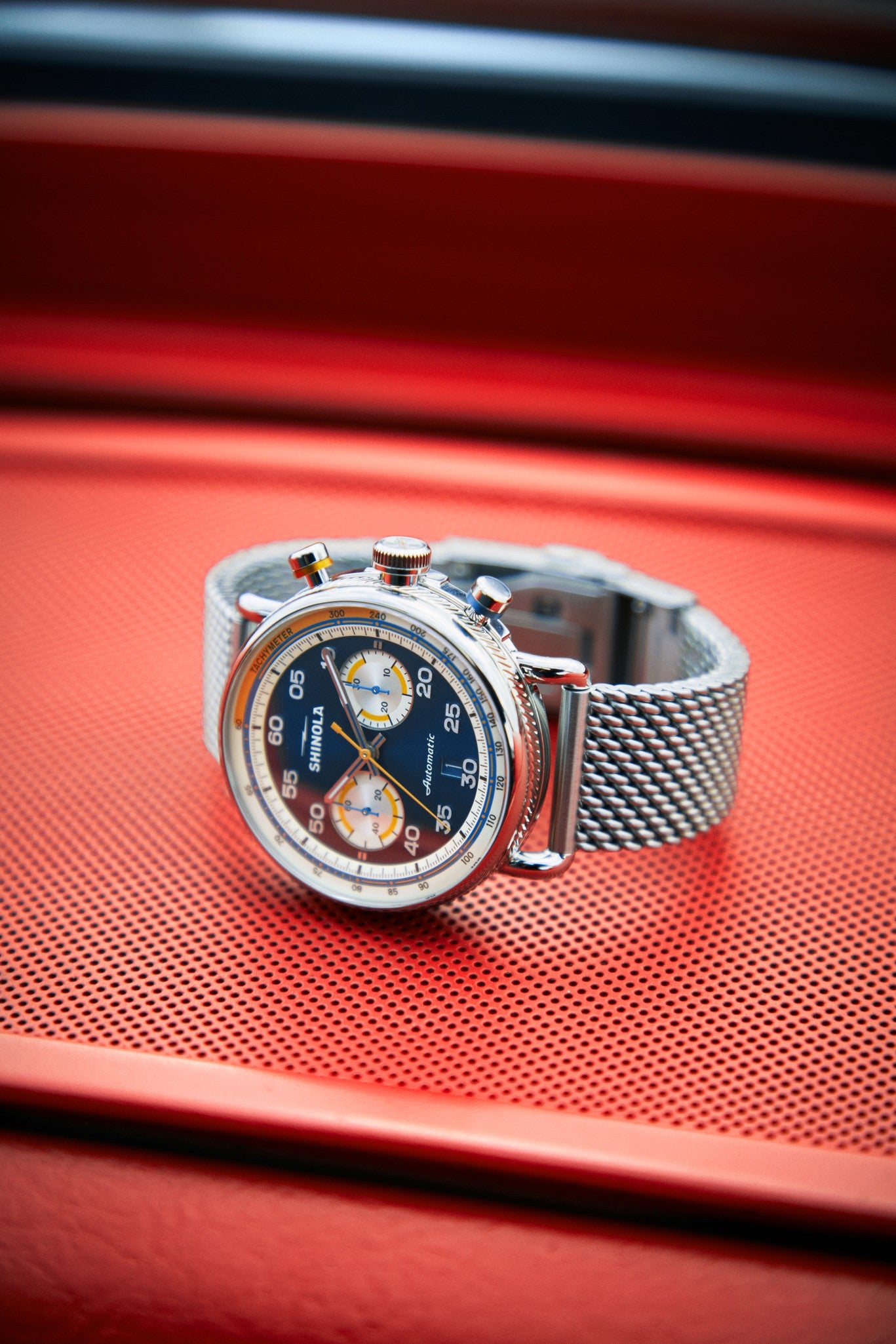 The Shinola Canfield Speedway Open Road Automatic Chronograph Watch - Blue, with a mesh band and a blue, white, and yellow chronograph dial featuring a tachymeter scale, is displayed on an orange perforated surface.