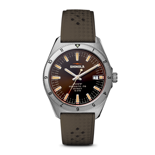 The Shinola Duck Watch - Mallard Brown features a brown gradient dial, silver hour markers, luminous hands, a silver bezel with black numerals, and a textured brown strap with Shinola and Detroit branding.
