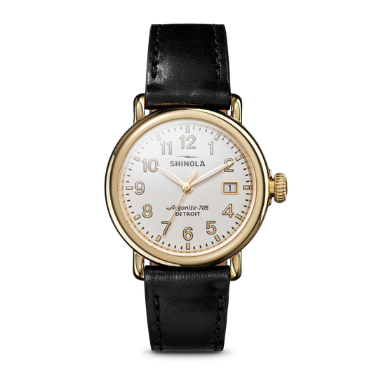 The Shinola Runwell Watch - Alabaster features a gold stainless steel case, white dial with black numerals, gold hands, date display, and a black leather strap, displayed on a plain white background.