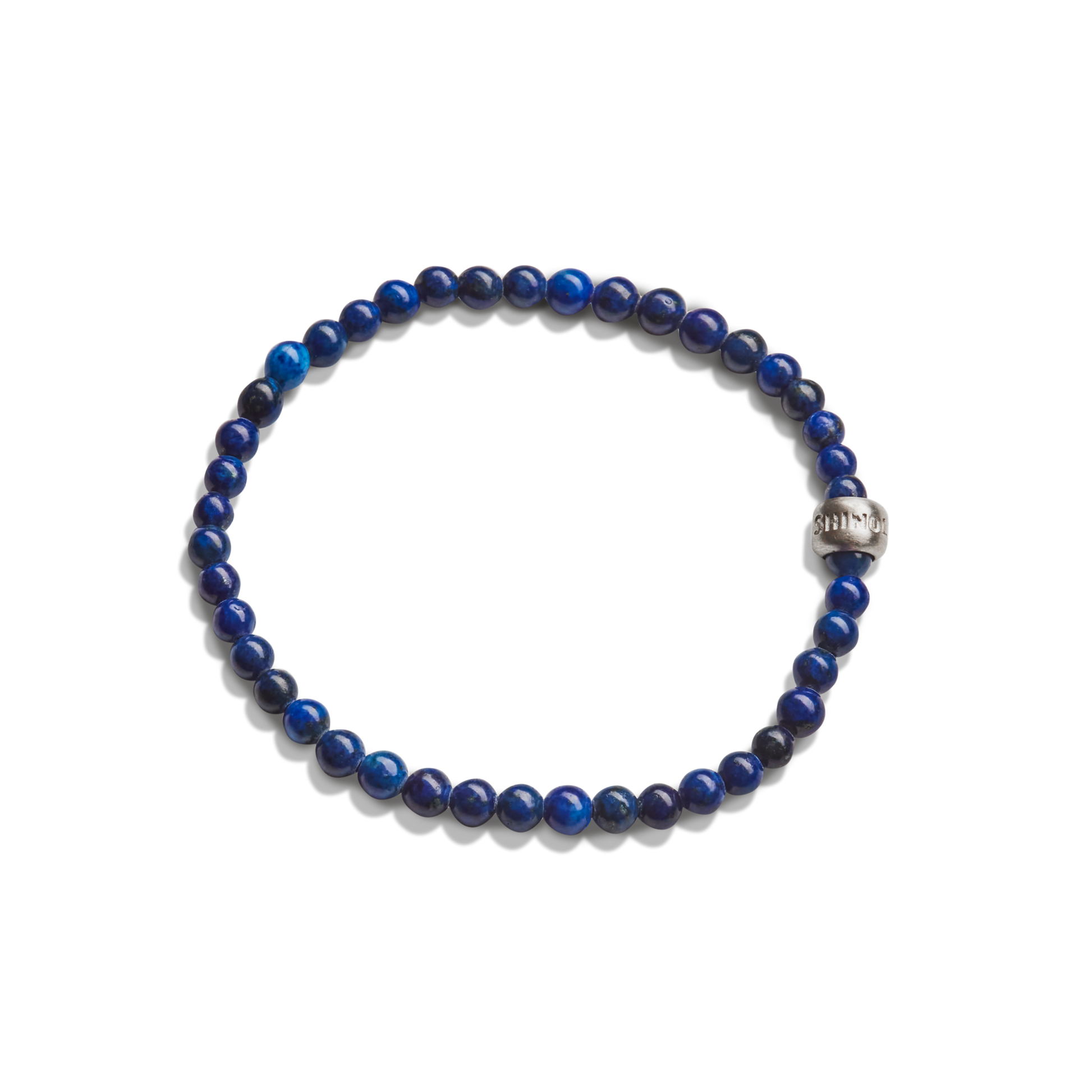 The Shinola 4mm Beaded Lapis Bracelet features round, dark blue lapis stones and a single engraved metallic bead, arranged in a circle against a white background.