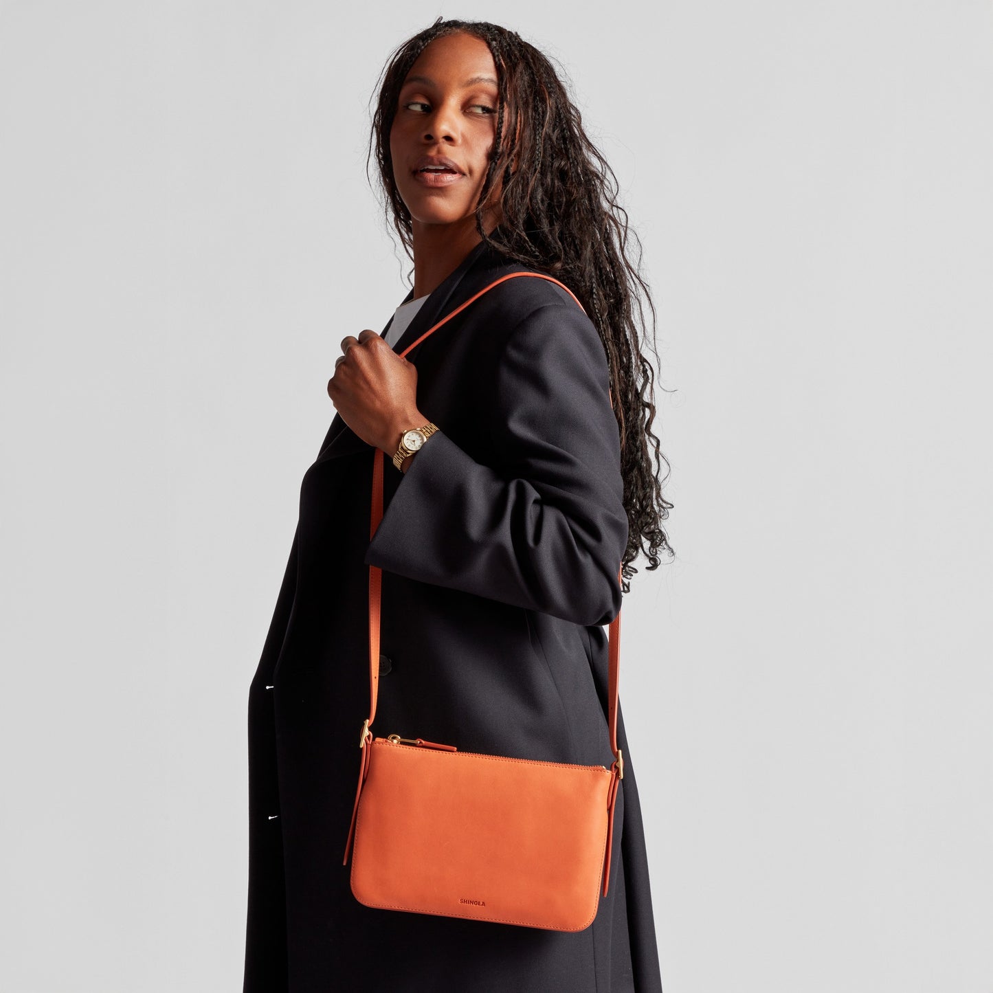 A woman with long braided hair wears a black coat and carries the Shinola Small Bixby Crossbody in Rust over her shoulder, looking off to the side against a plain light background—an effortless everyday accessory.