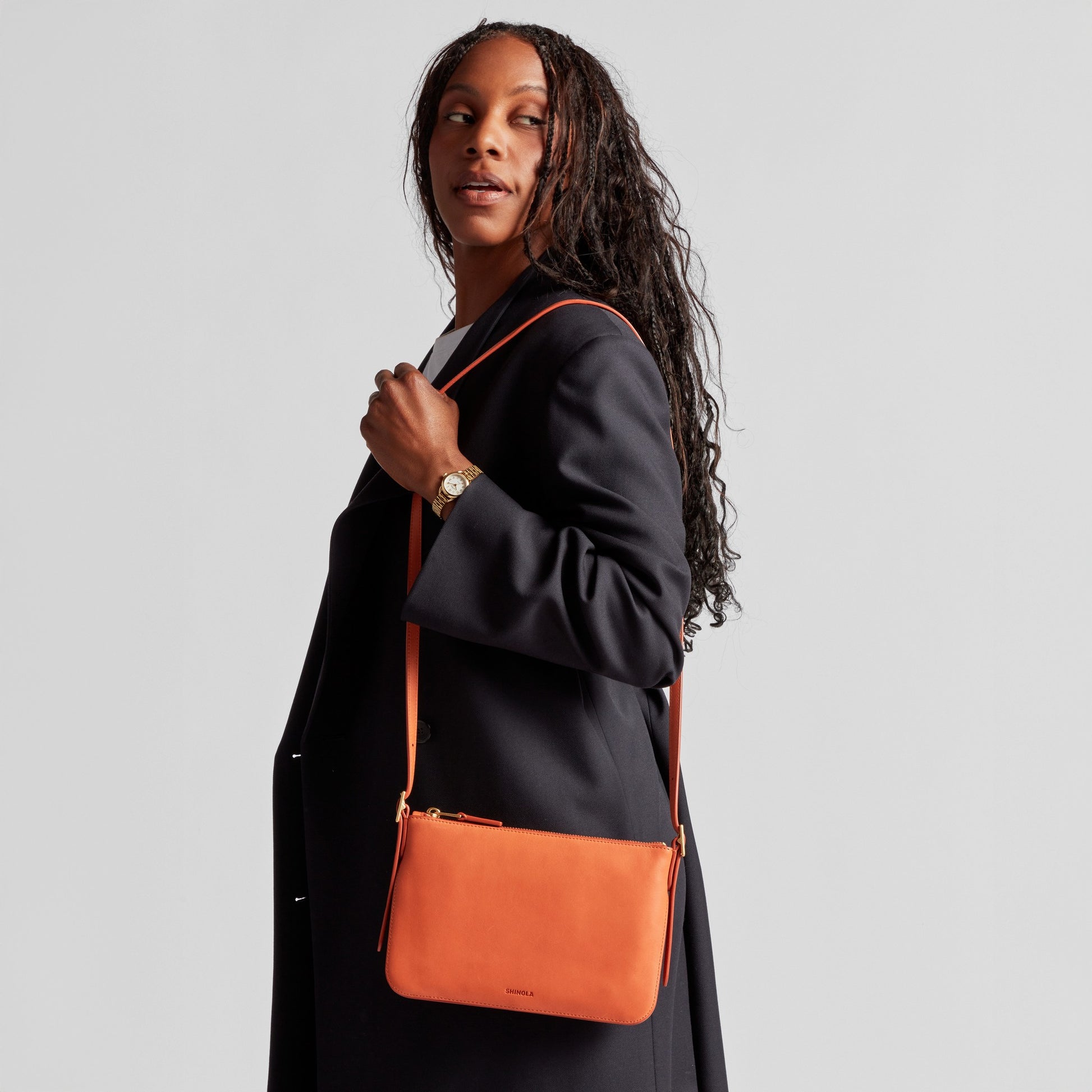 A woman with long braided hair wears a black coat and carries the Shinola Small Bixby Crossbody in Rust over her shoulder, looking off to the side against a plain light background—an effortless everyday accessory.