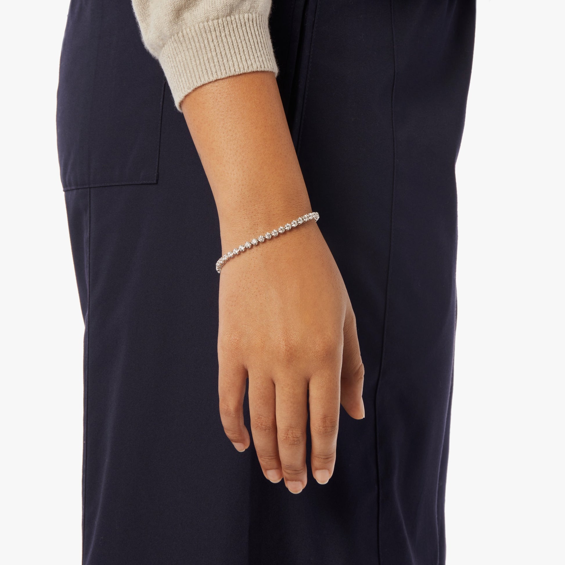 A person is shown from the waist down wearing dark pants and a light top. On their wrist is the Shinola 4 Carat 7" Diamond Tennis Bracelet in White Gold, featuring brilliant cut diamonds.