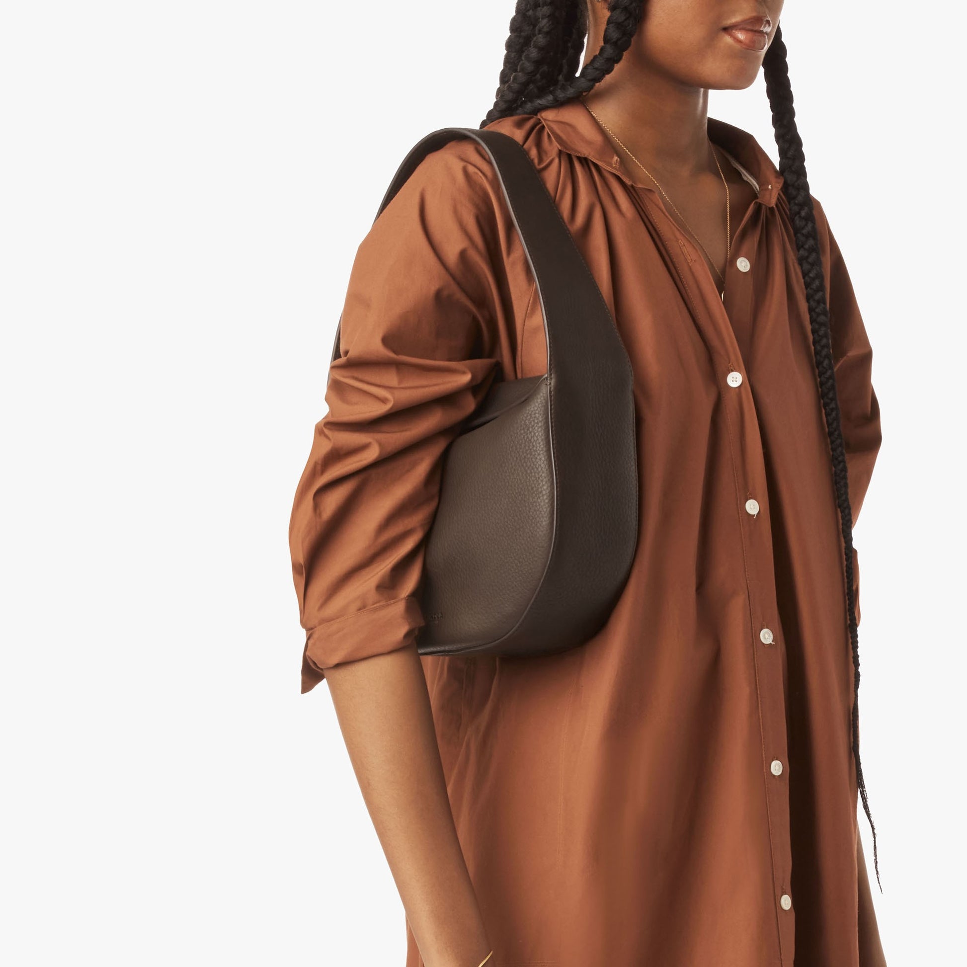A person in a brown long-sleeve button-down dress holds the Shinola Small Shoulder Bag - Espresso, crafted from natural grain leather. Part of their face is visible, with long braided hair, against a white background.