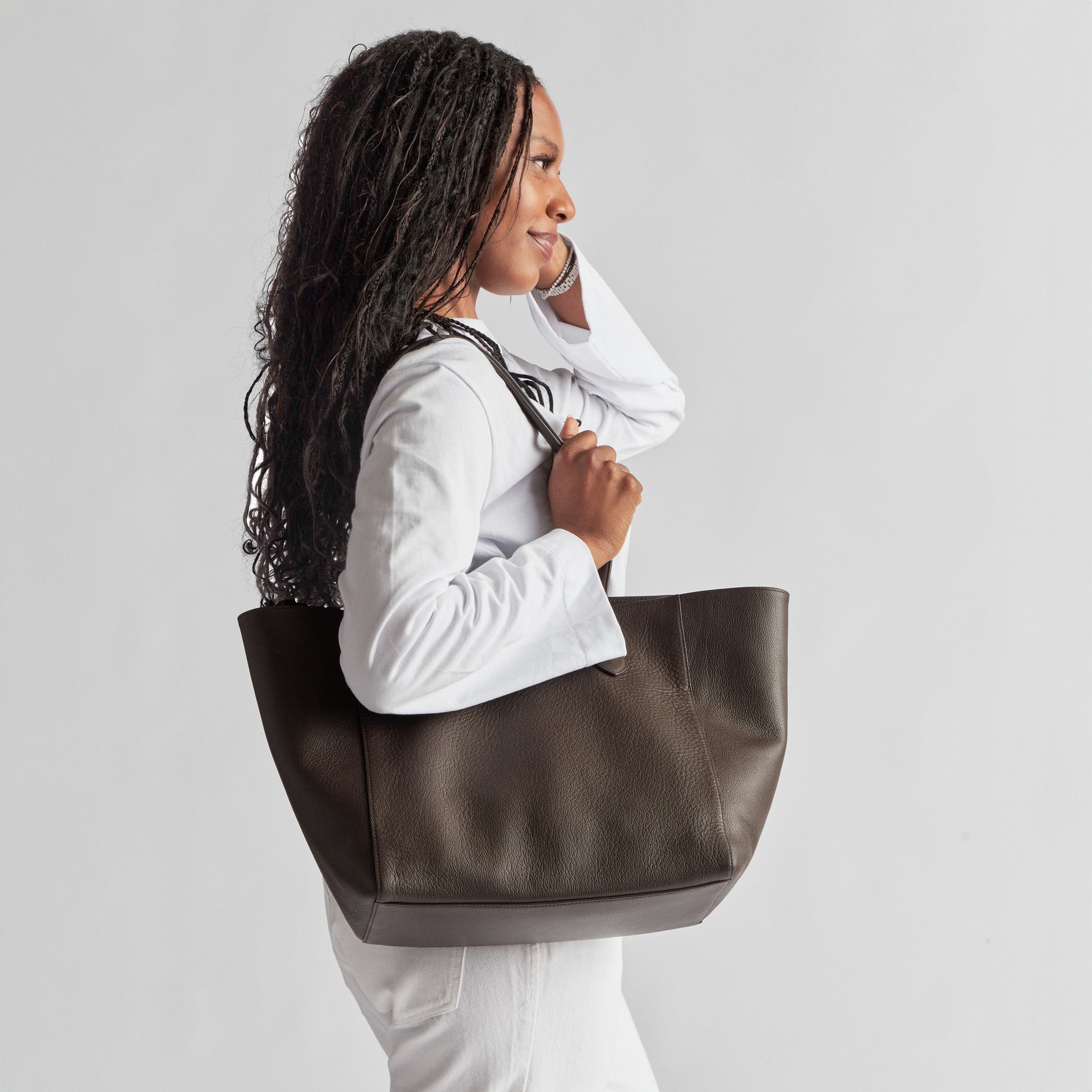 A woman with long braided hair in a white outfit stands in profile, carrying the Shinola Runabout Tote Bag - Espresso, a large, lightweight pebbled leather accessory, over her shoulder against a plain light background.