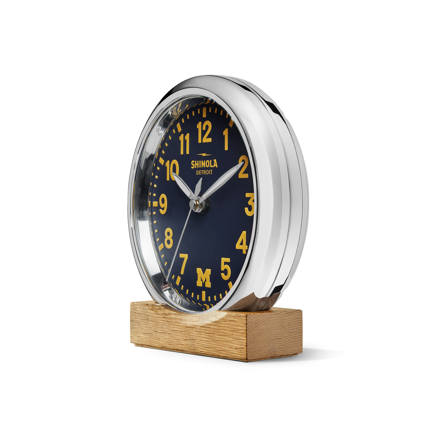 A 6" Runwell Clock by Shinola features a wolverine blue face with yellow numbers, hands, and "M" logo, celebrating the University of Michigan. It has a chrome rim, sits on a wooden base, and reads "Shinola Detroit" on the face.