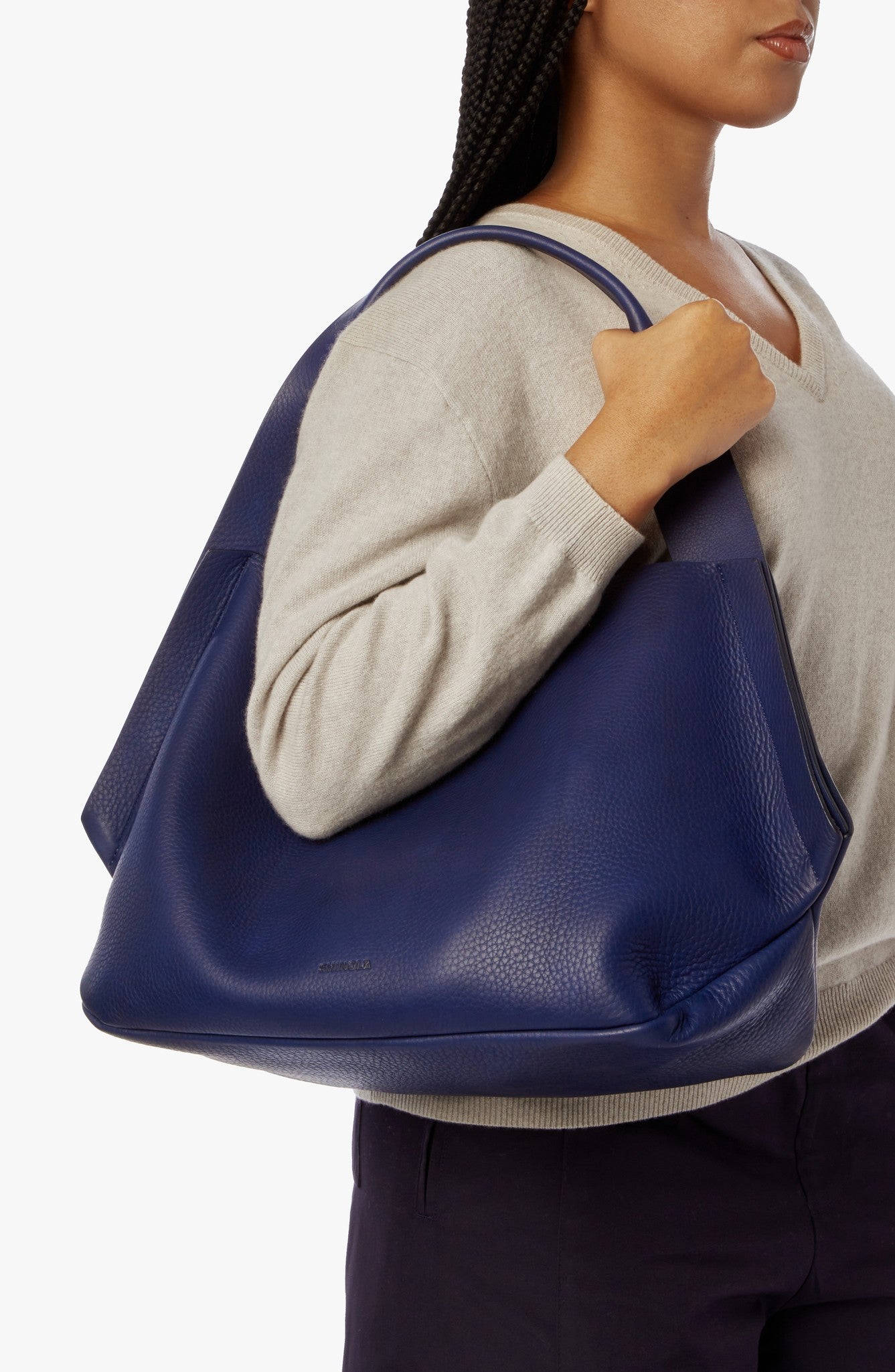 A woman in a light beige sweater holds the Shinola Runwell Shoulder Bag - Lake, a large, deep blue leather bag with wide handles and a convenient leather card pocket. The bag has a smooth, slightly textured finish; the background is plain and light.