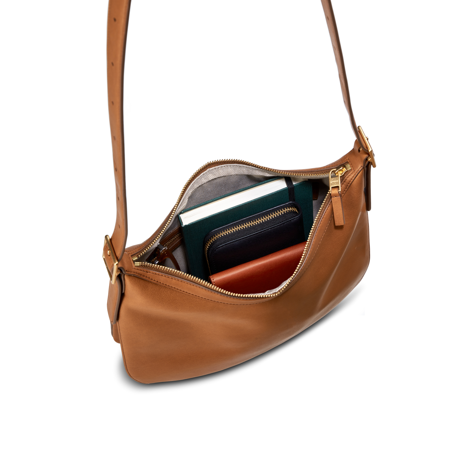 The Shinola Medium Bixby Crossbody in tan natural leather is a versatile accessory, shown with its zipper partially open to reveal a green notebook, black wallet, and brown item neatly organized inside.