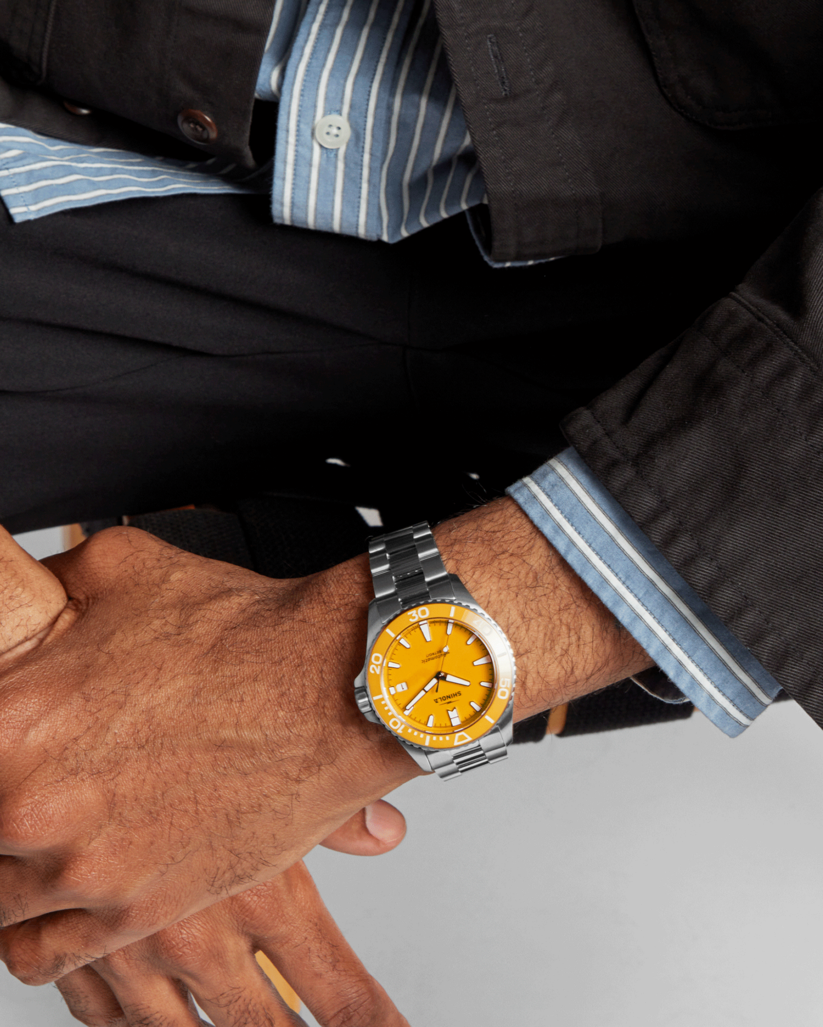 A person in a dark jacket and blue striped shirt rests their hands on their leg, showcasing a Shinola Lake Huron Monster Automatic Dive Watch with a stainless steel case and bright orange face.