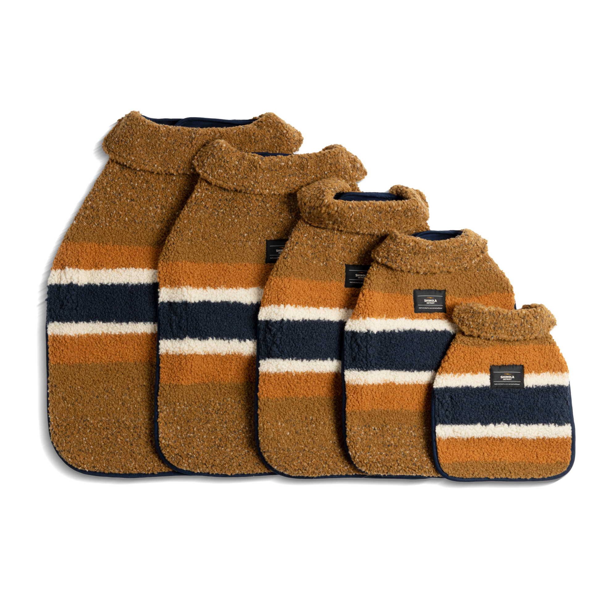 Five tan Shinola Dog Coats with white, orange, and navy blue stripes are arranged by size against a white background. Each premium Brand Stripe coat features a soft collar and a small dark label on the front.