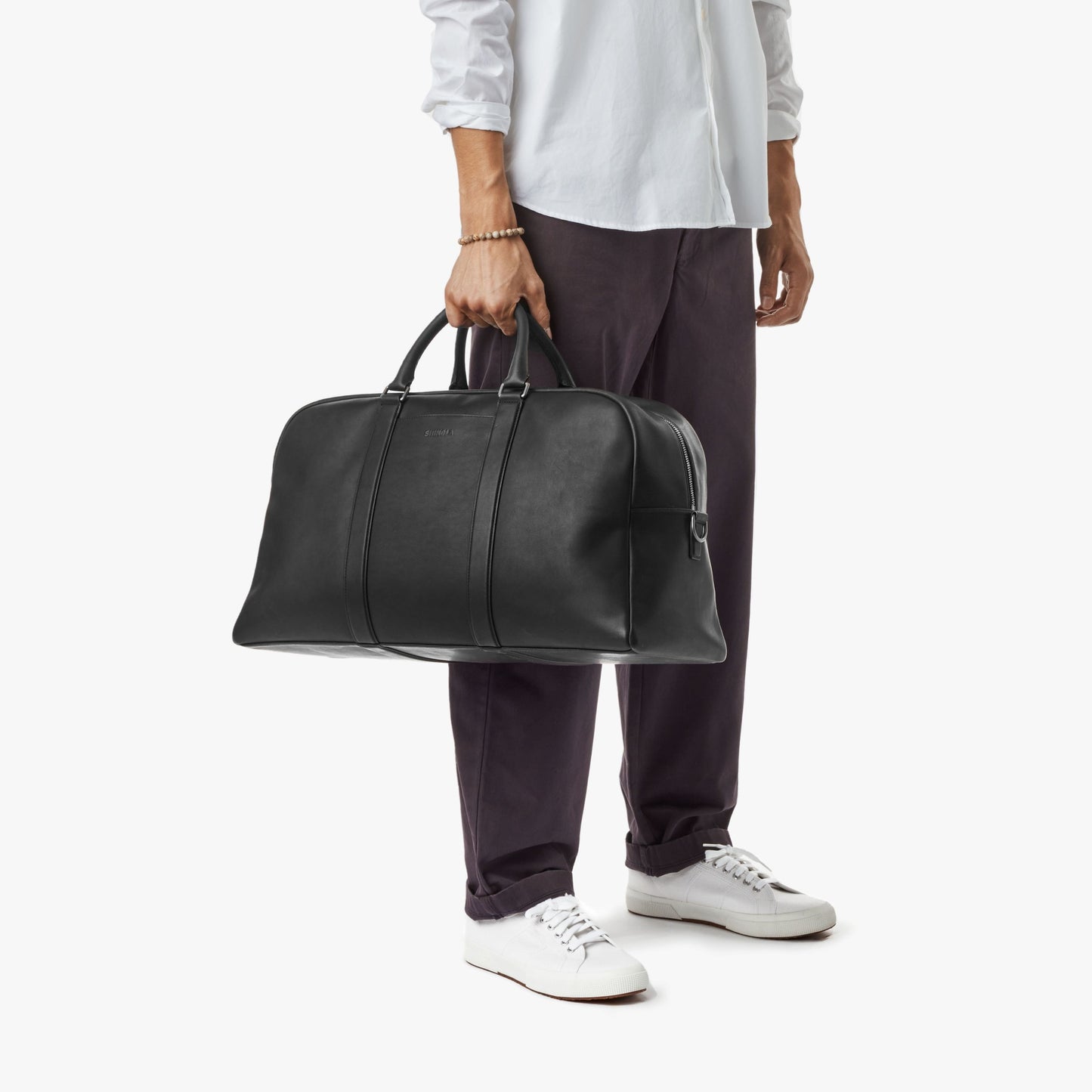 A person in a white shirt, dark pants, and white sneakers holds the Shinola Runwell Duffle Bag - Black by its handles against a plain white background.