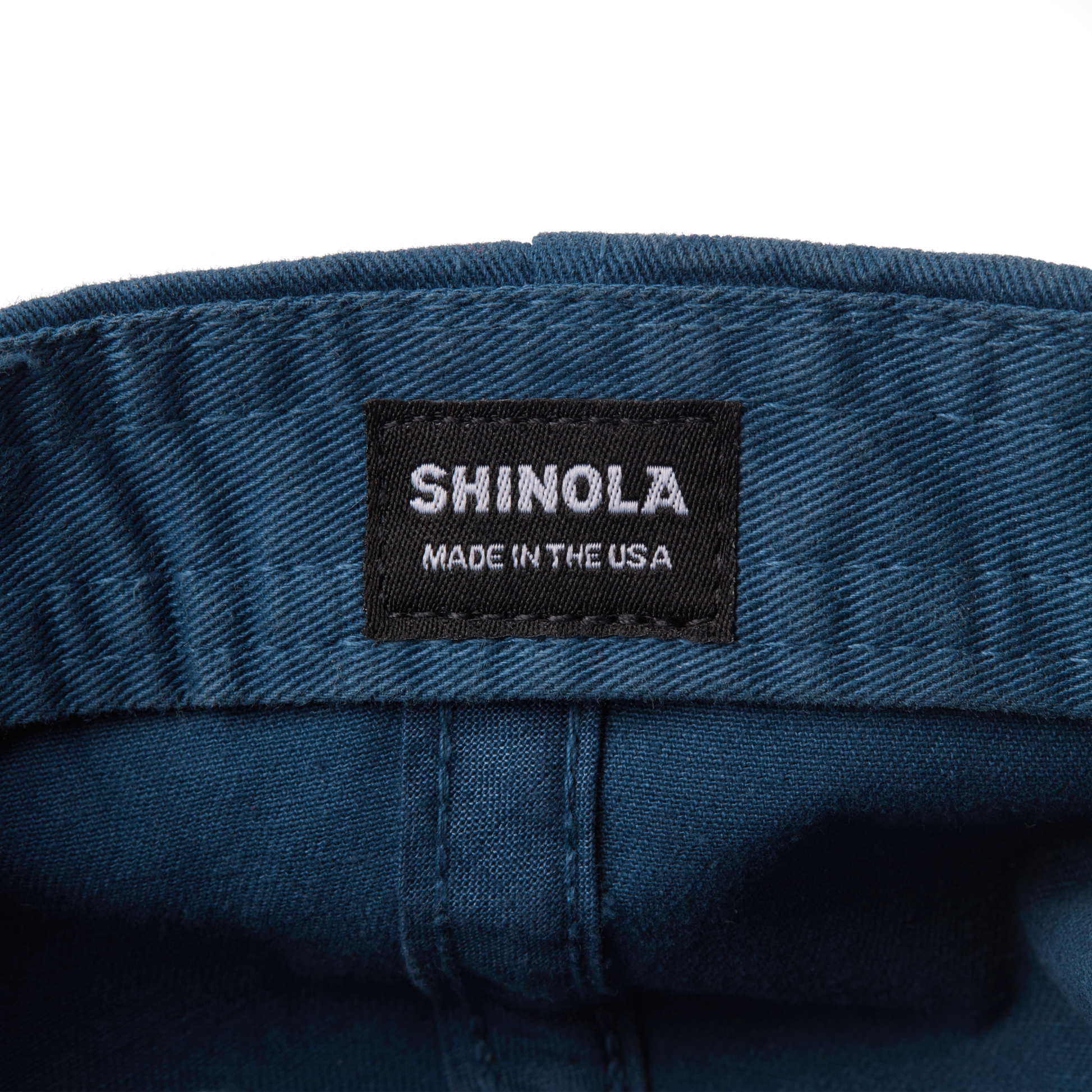 A close-up of a black fabric label sewn onto the blue Cotton Twill 6 Panel Cap by Shinola, displaying the text SHINOLA MADE IN THE USA in white capital letters.