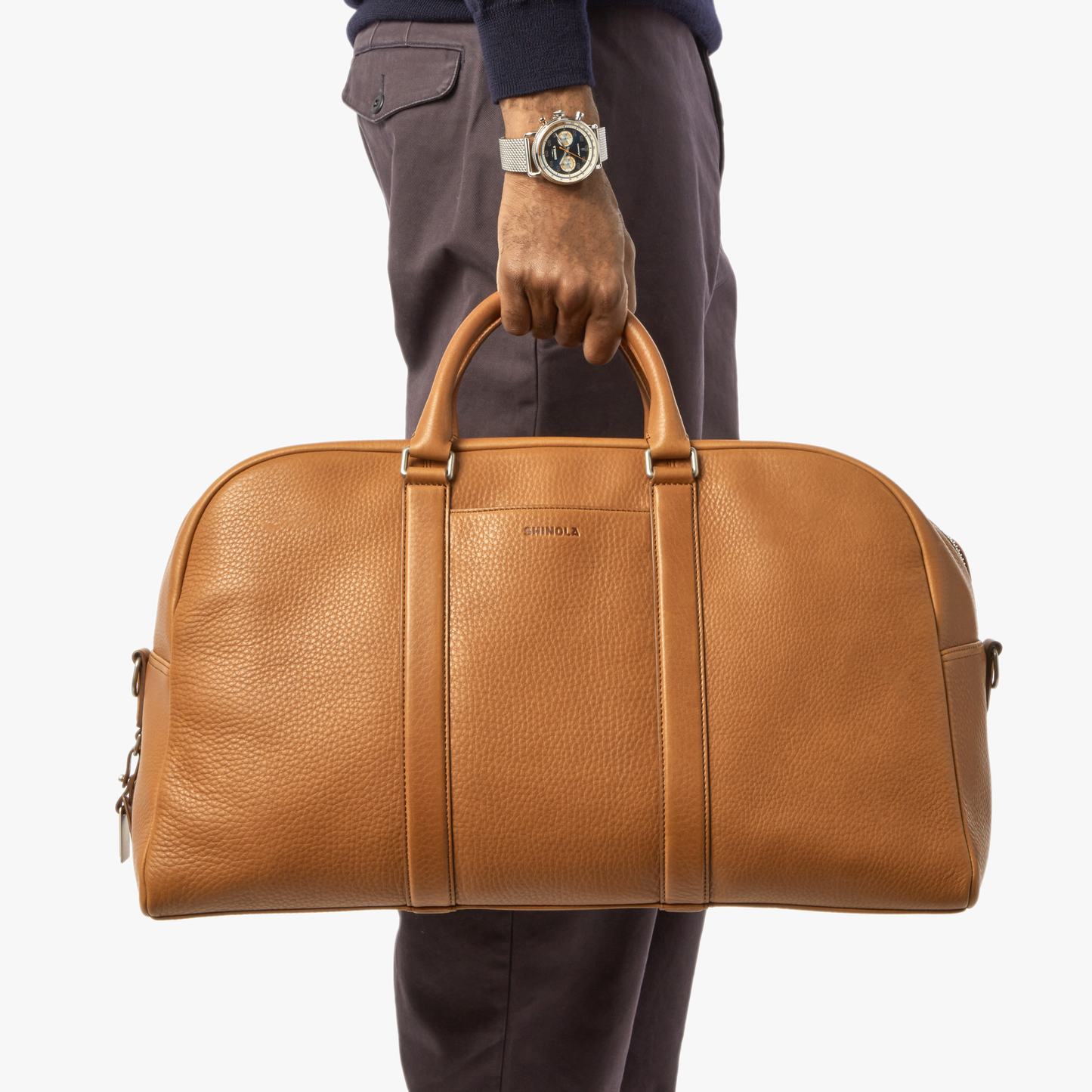 A person in brown pants and a watch holds the Shinola Runwell Duffle Bag - Tan, featuring two handles and ideal as a TSA carry-on, against a white background.