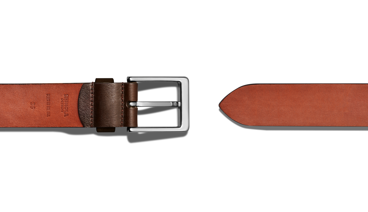 The Shinola 1 1/2" Lightning Bolt Keeper Belt in Deep Brown, featuring silver-tone hardware and a rectangular buckle, is shown laid flat on a white background. This casual belt includes a pointed tip and embossed inner text for detail.