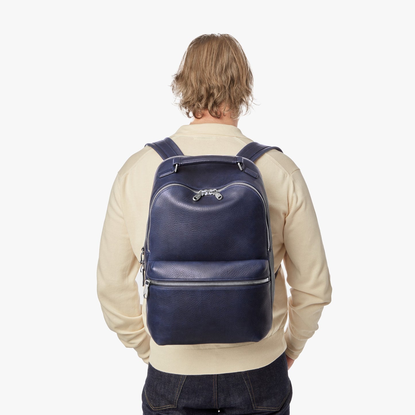 A person with blonde hair, in a light beige sweater and dark jeans, is shown from the back carrying the Shinola Portfolio Briefcase - Stone against a plain white background.